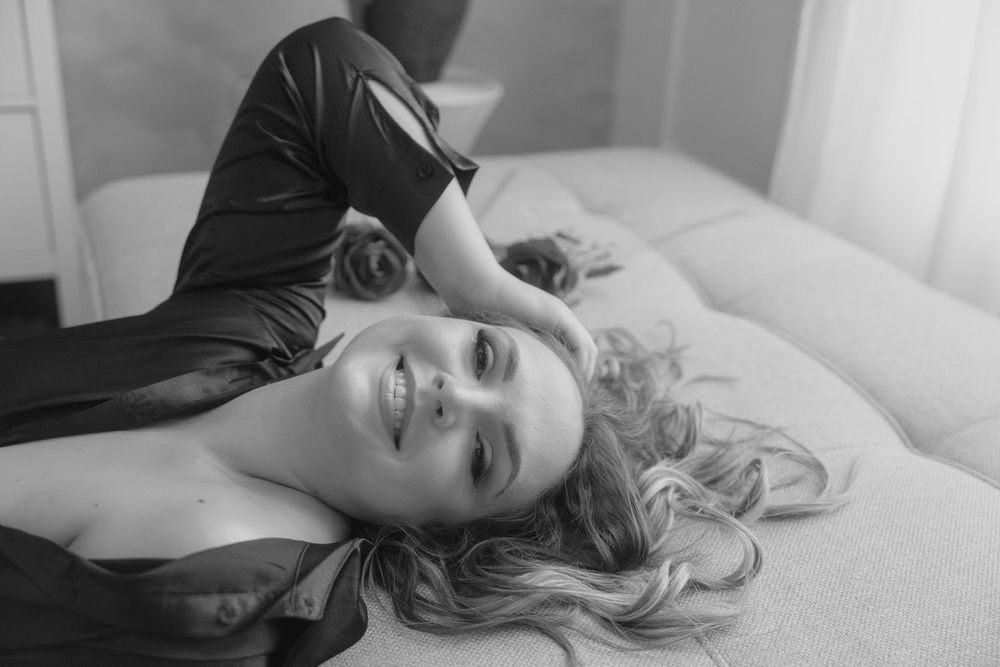 Black and white portrait of someone in dark clothing lying on white bedding.