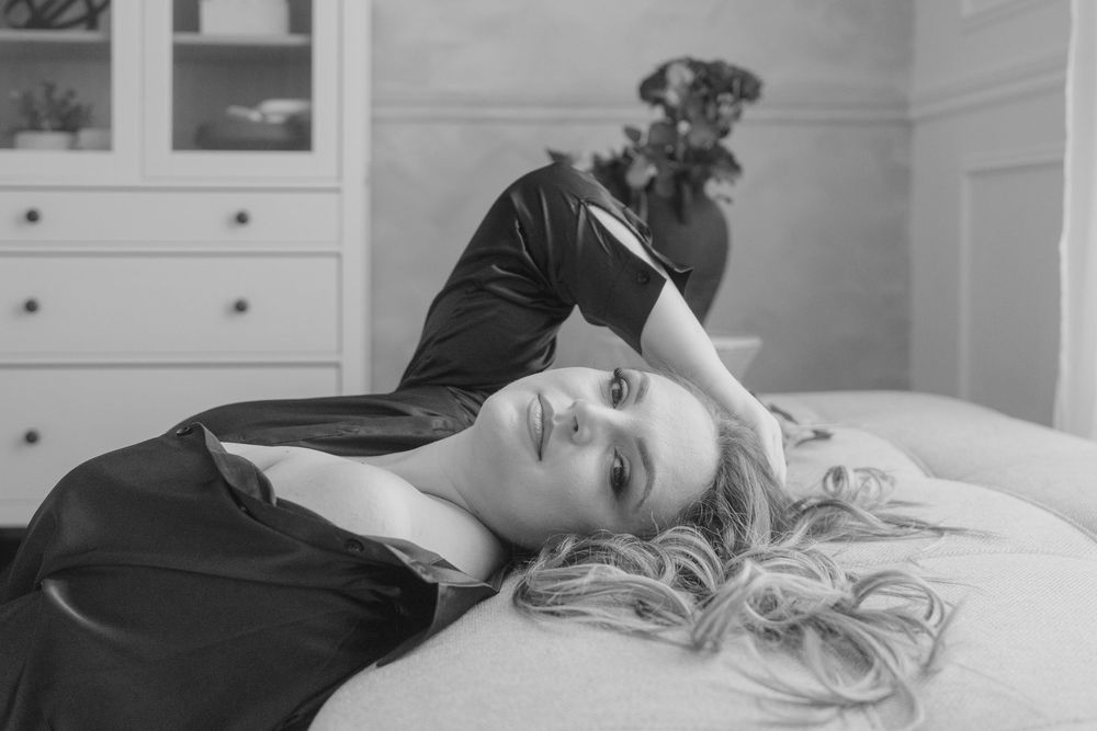 Artistic black and white boudoir photograph showing relaxed pose on bed with white linens.