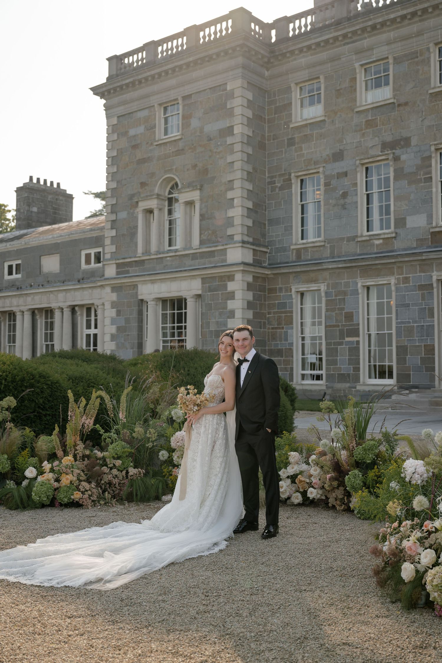 A wedding couple poses in front of a grand stone manor house with elegant floral arrangements on a gravel path.