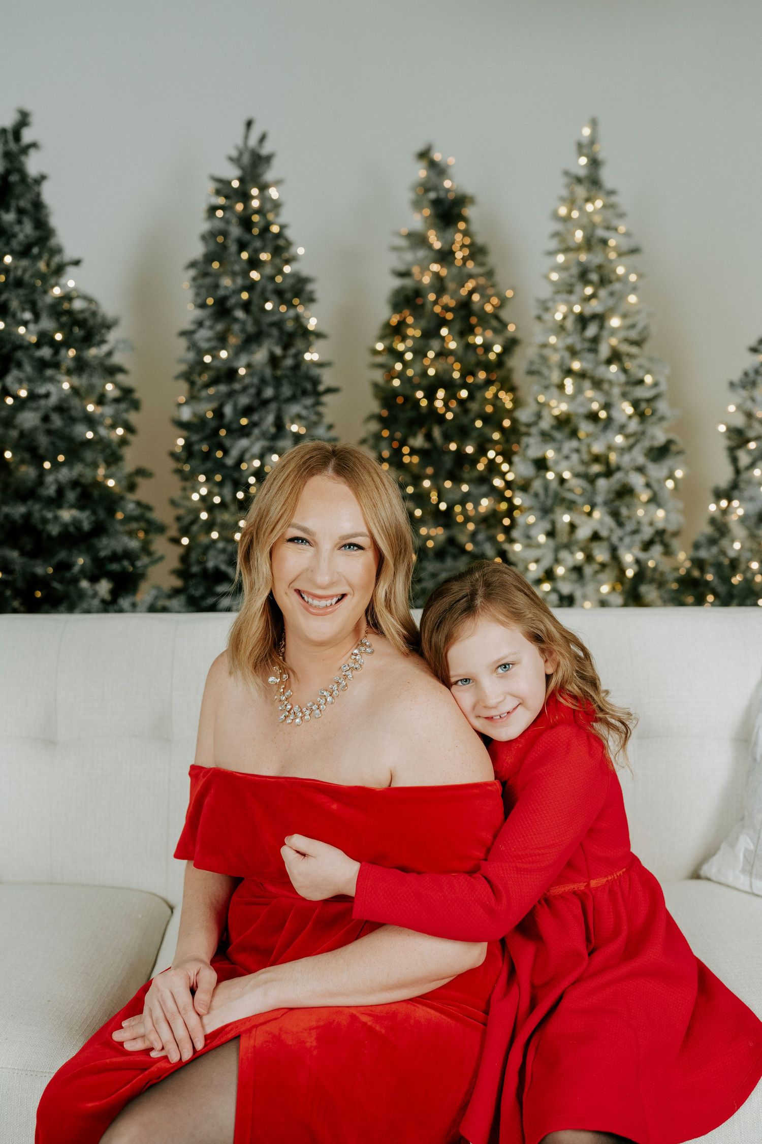 Mother and daughter in matching red dresses pose for holiday photos in front of decorated Christmas trees.