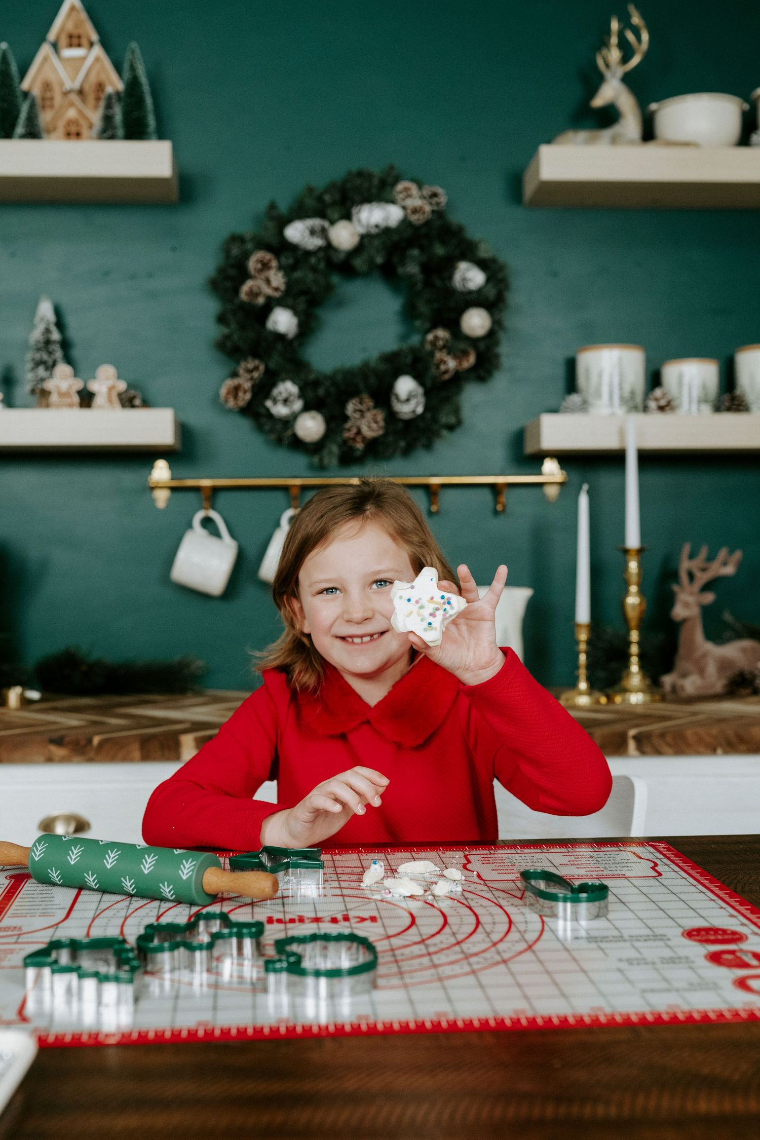A child wearing red sits at a table with holiday baking supplies and cookie cutters.