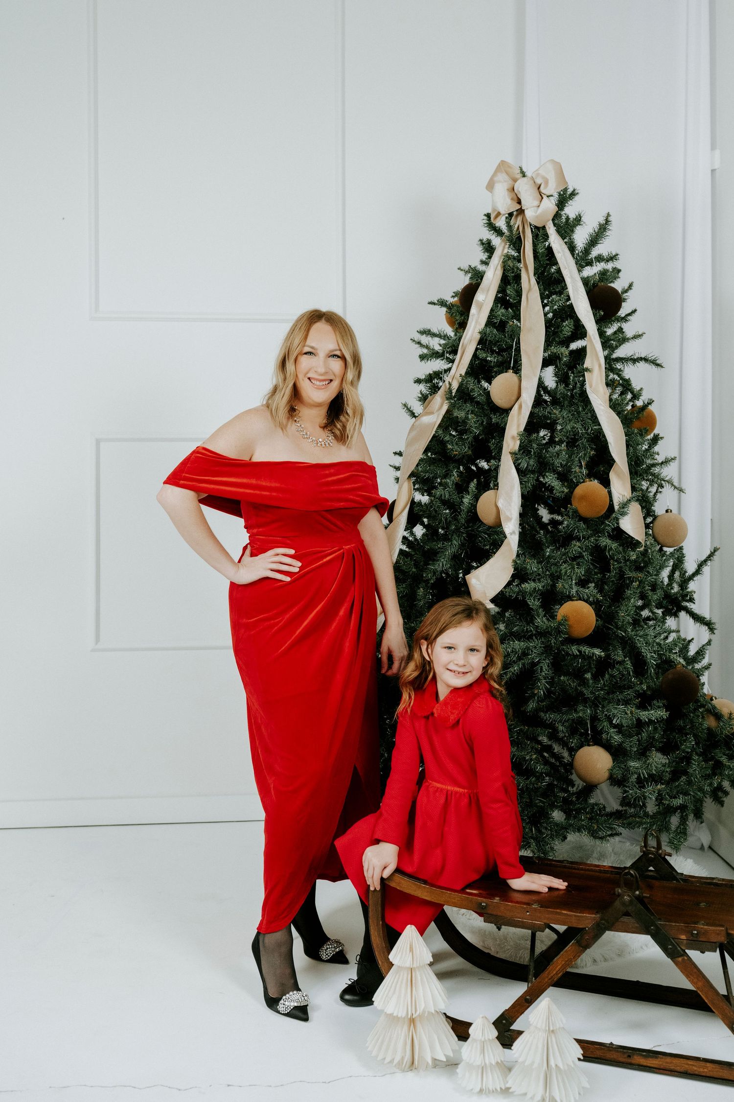 Matching red holiday outfits create a festive scene with Christmas trees and decorative sleigh in background.