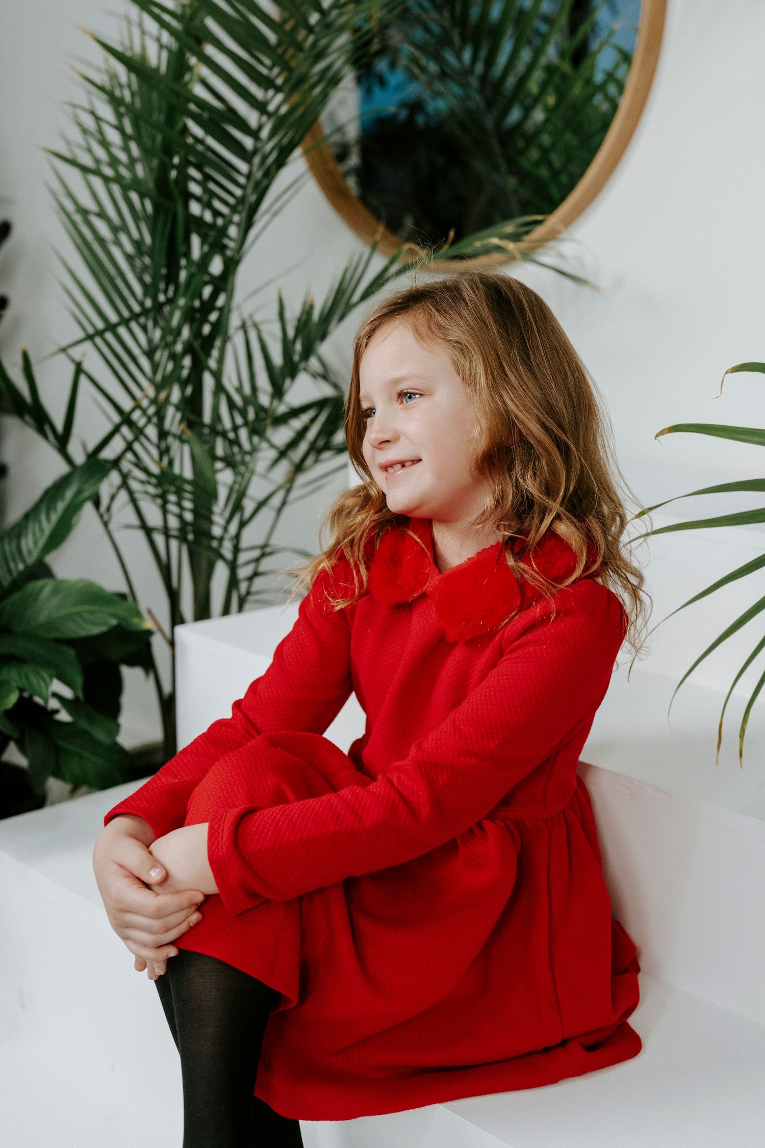 A child in a red sweater smiles and poses against a white wall with tropical plants in the background.