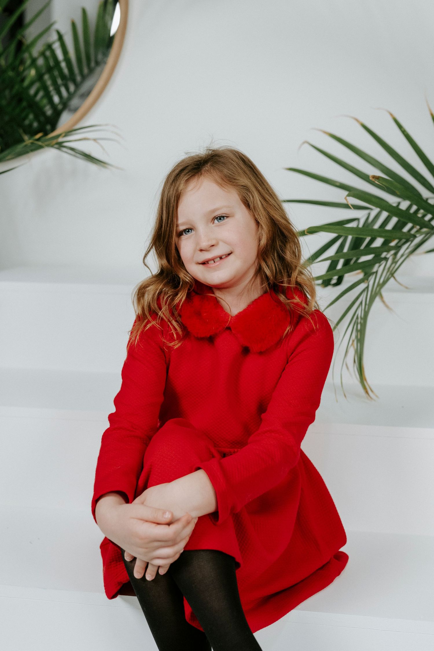Child in bright red jacket sits on white steps with tropical palm leaves in background.
