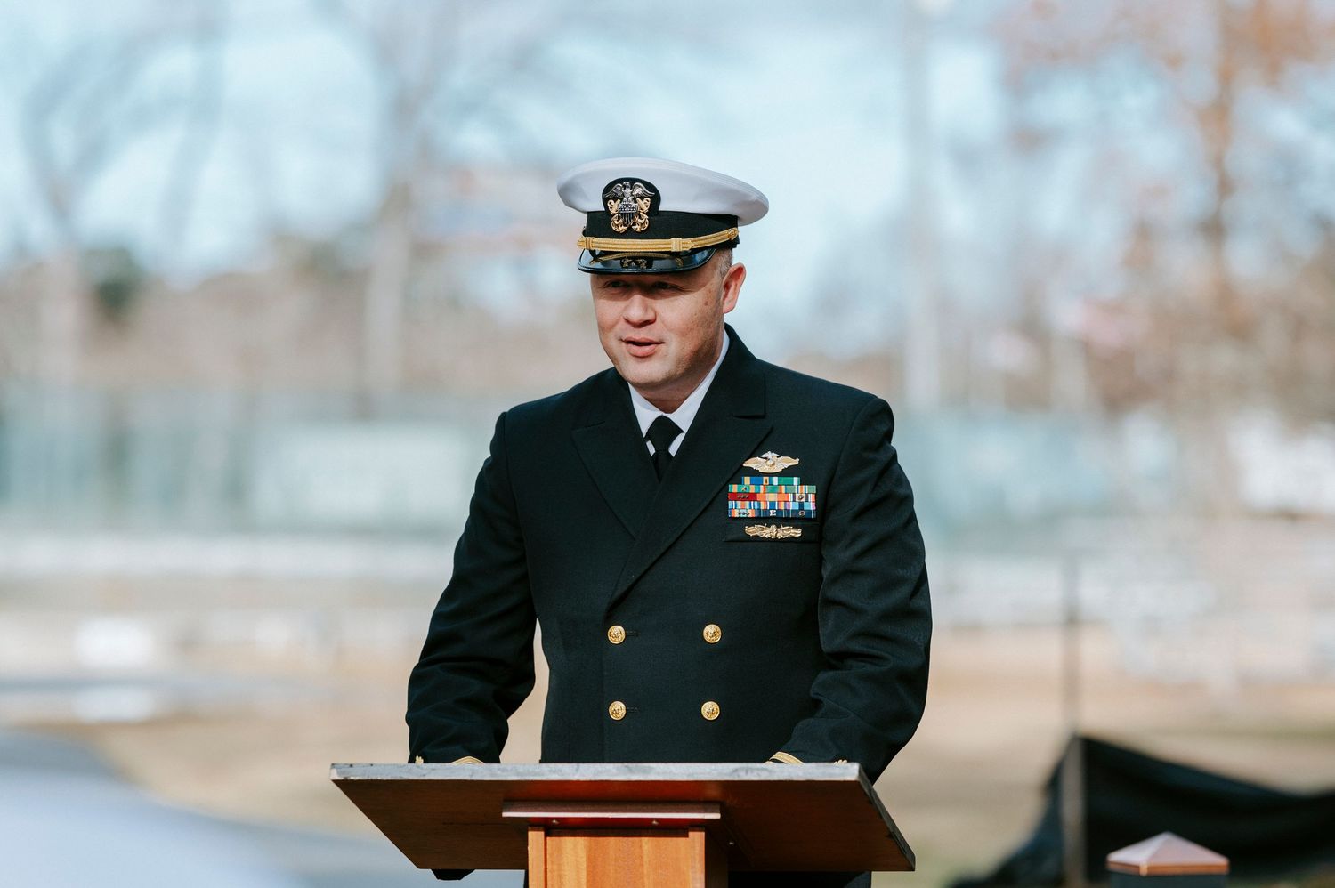 Military officer in dress uniform stands at podium during outdoor ceremony in autumn setting.