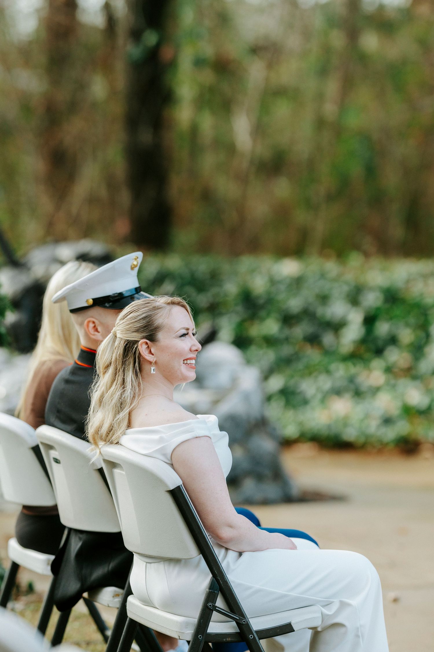 A person in a white dress sits in a white chair outdoors while looking to the side with greenery in the background.
