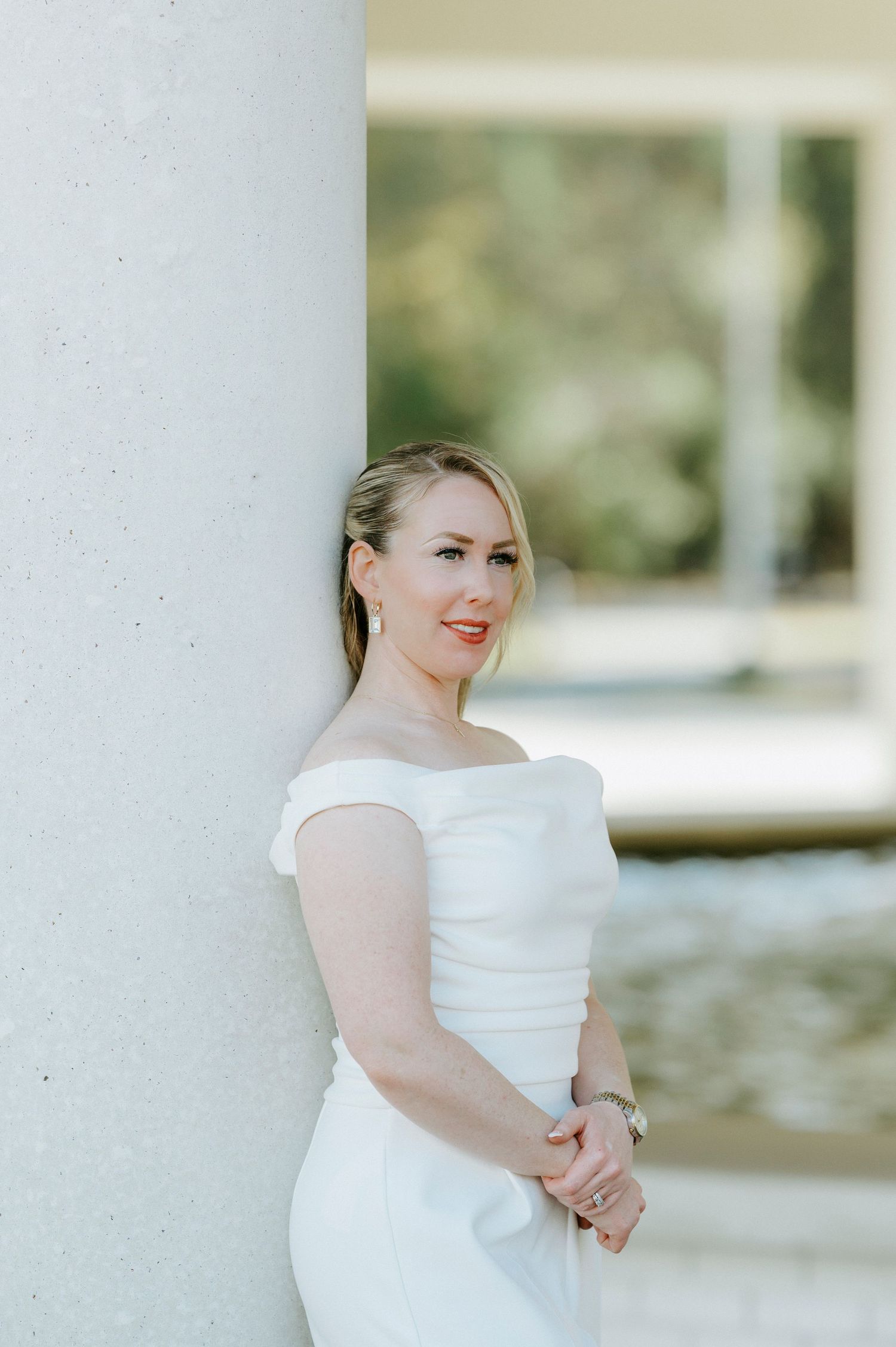 Elegant bride in a fitted off-shoulder white wedding dress poses against a white pillar in natural light.
