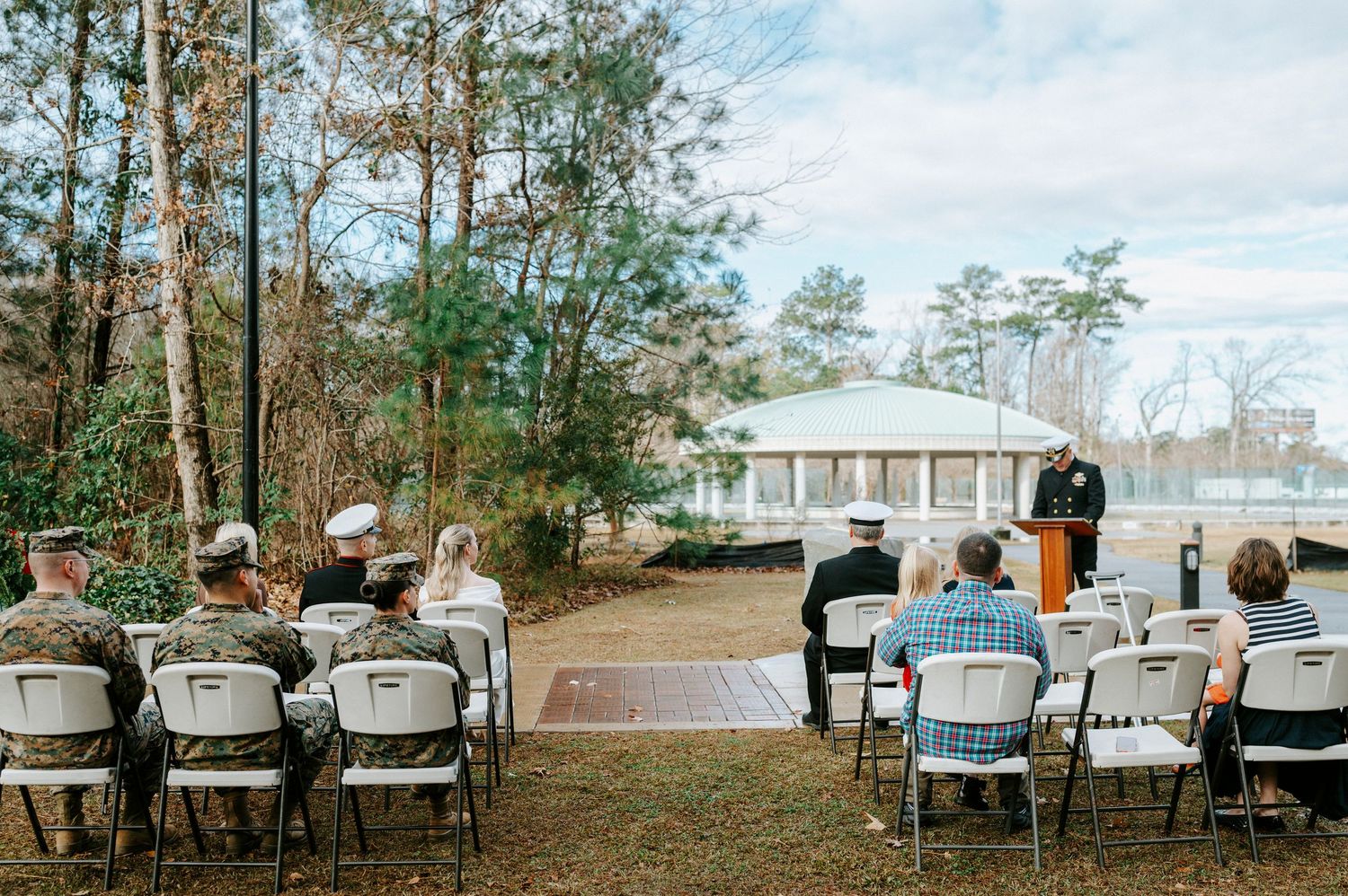 White chairs arranged in rows for an outdoor ceremony against a scenic wooded backdrop with a white gazebo in the distance.