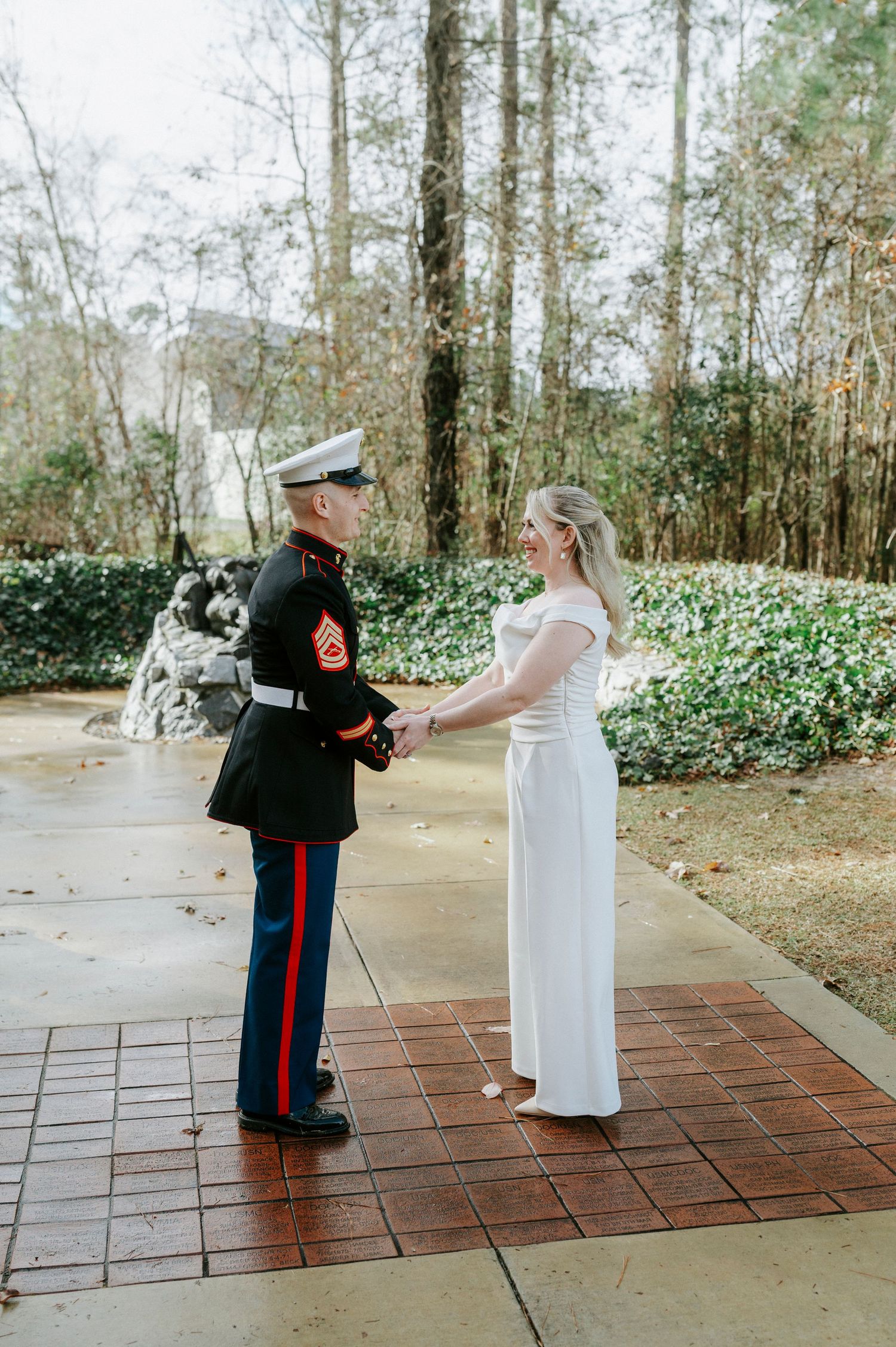 Military groom in dress uniform and bride in white gown share a moment on brick pathway surrounded by trees.