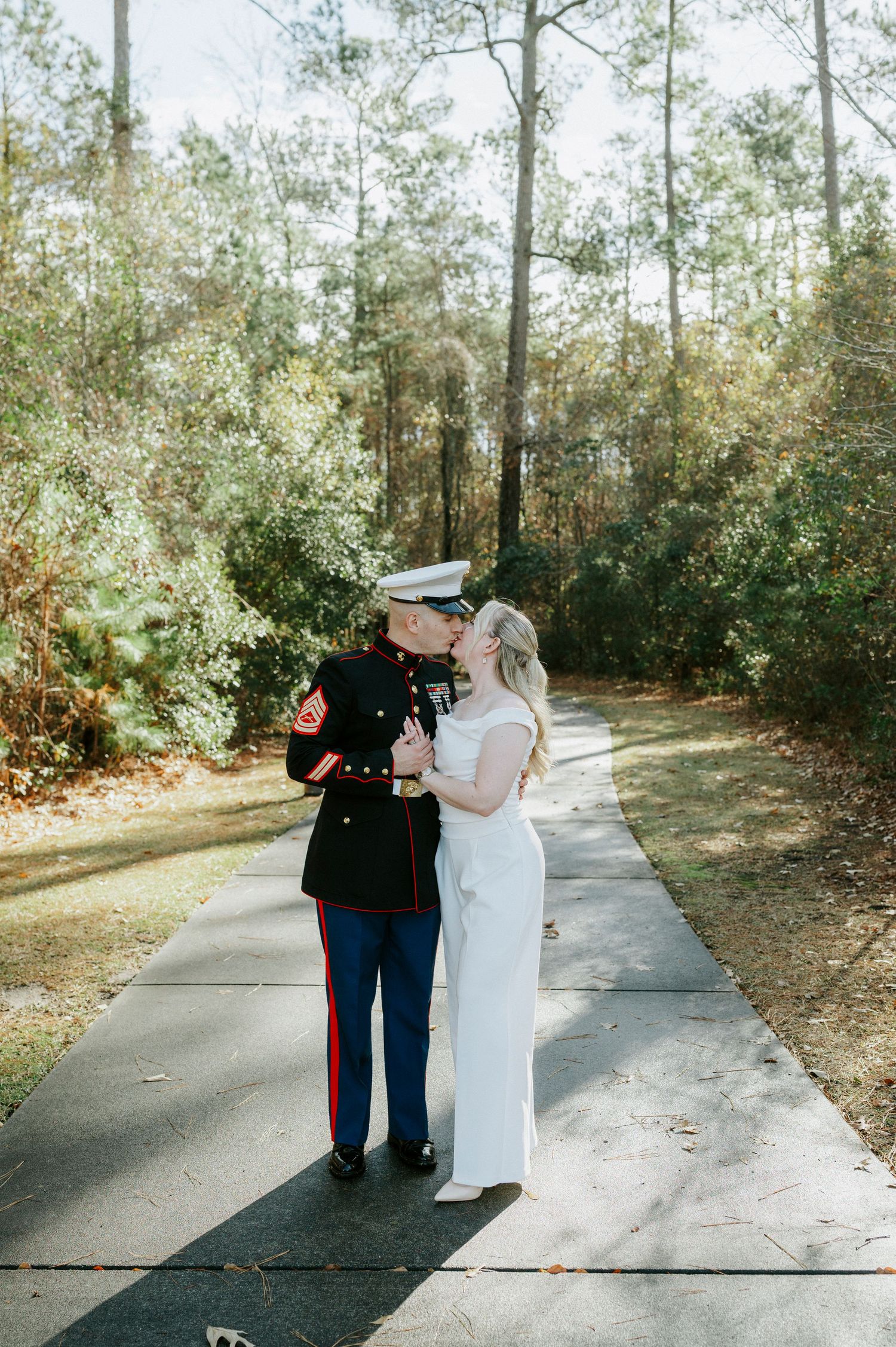 Military groom and bride in white dress walking together on sunlit path through wooded area.