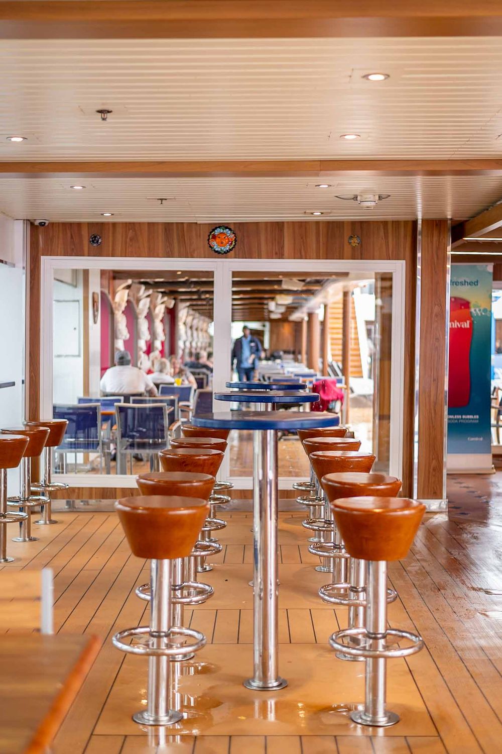 Modern cruise ship bar area with wooden deck flooring and sleek chrome bar stools arranged in rows.