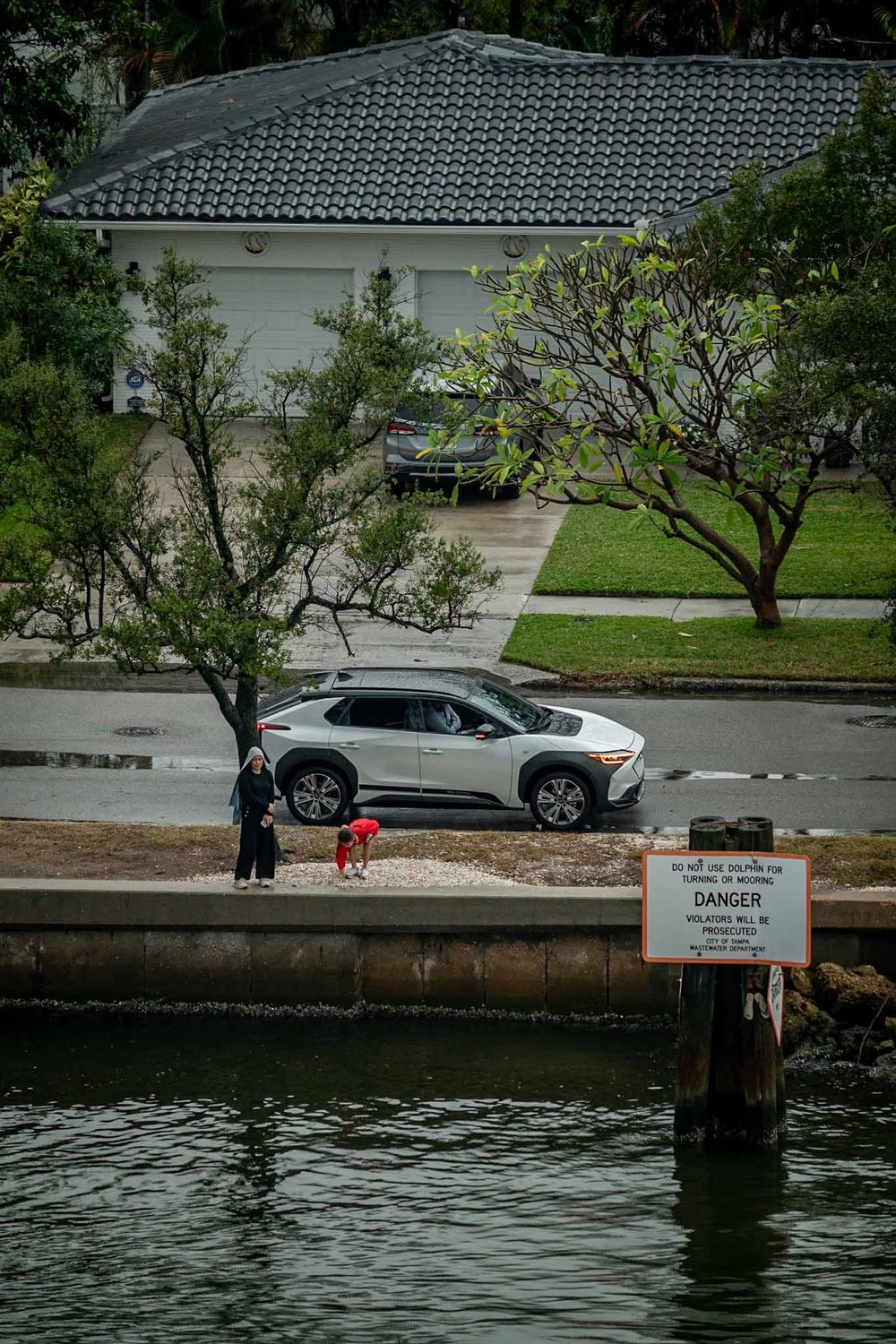 Warning sign posted along a waterfront walkway with parked cars and trees in the background.
