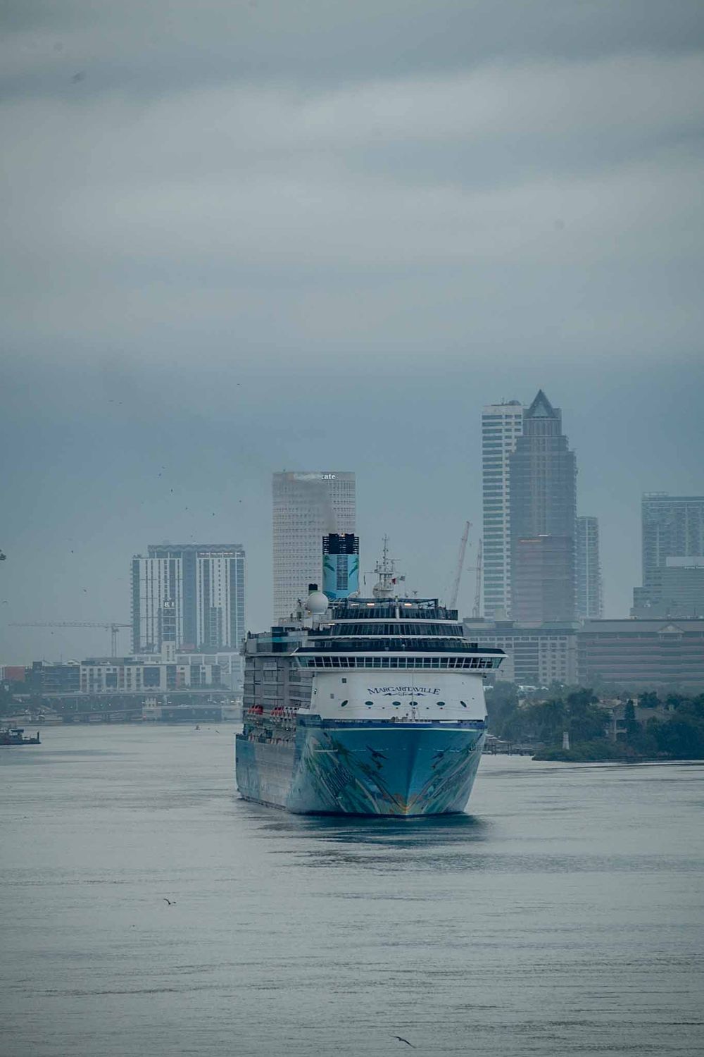 Three large cruise ships sailing in front of a misty city skyline on a cloudy day.