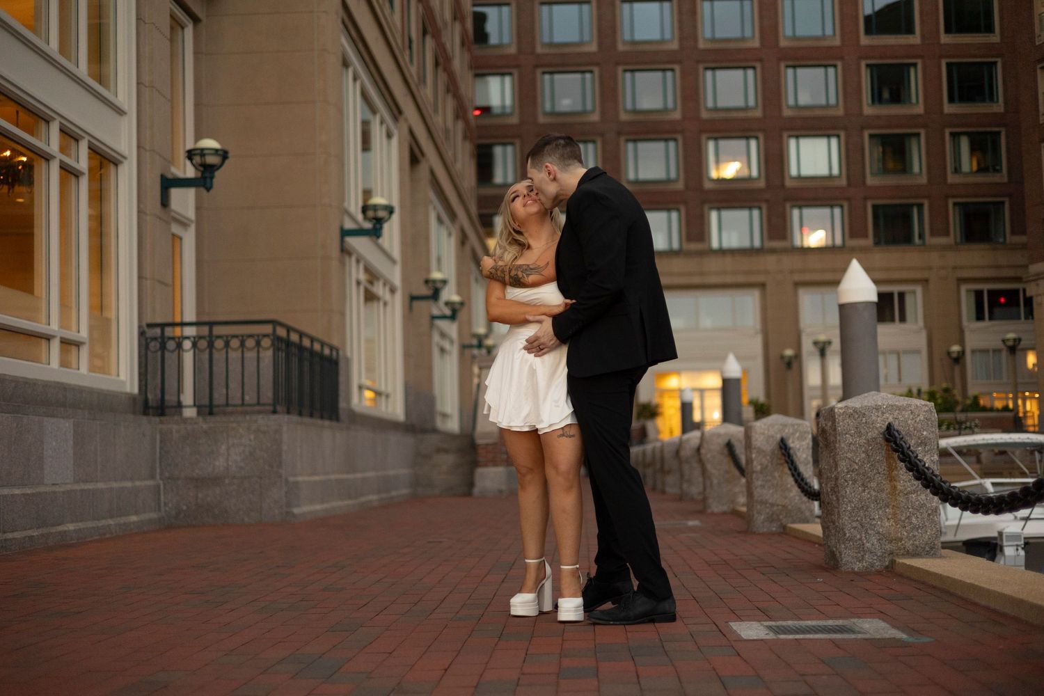 A couple shares an intimate moment on a brick terrace outside an urban building at dusk.