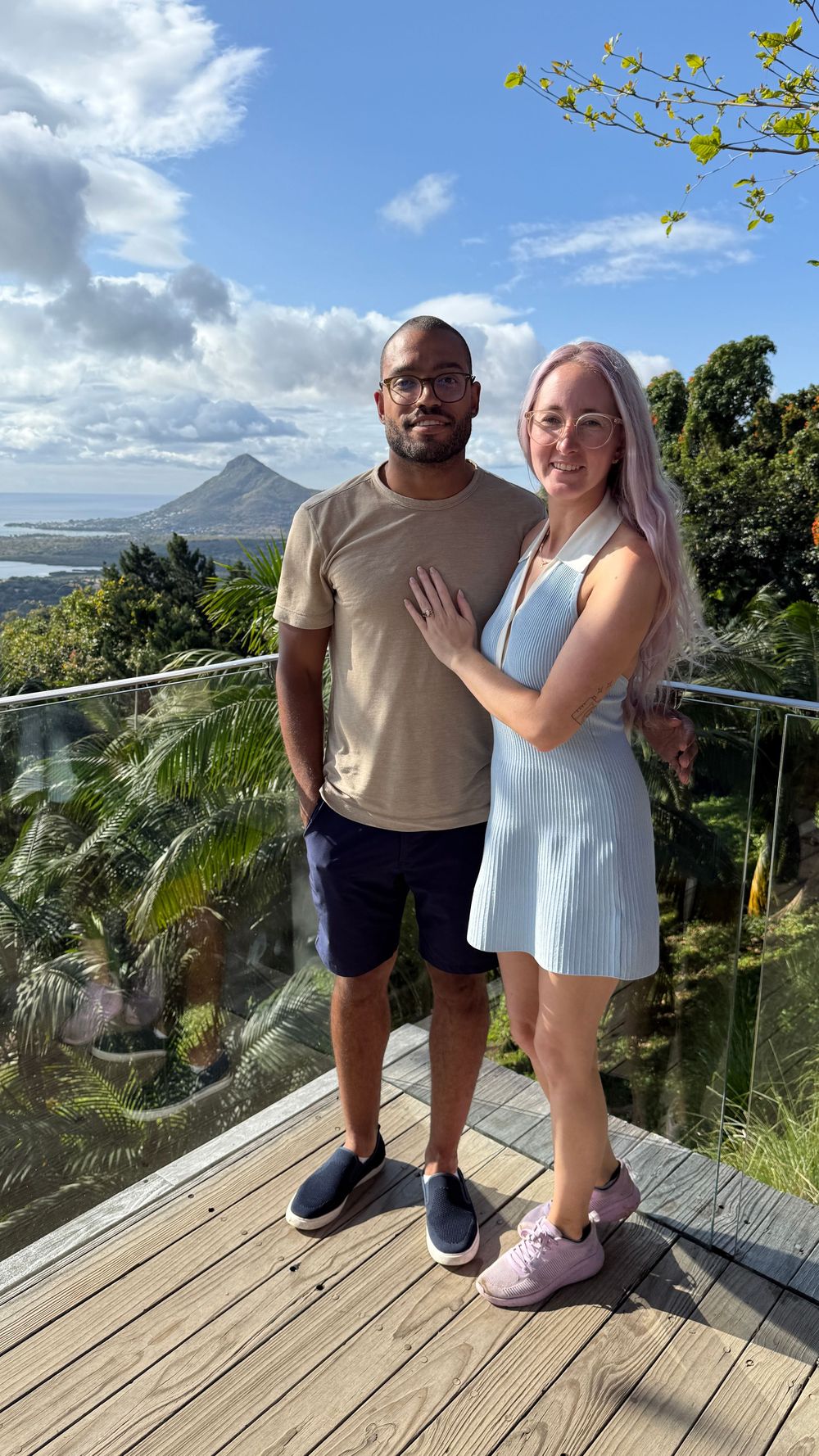 A couple poses together on a wooden deck overlooking a tropical mountain landscape with palm trees and blue skies.