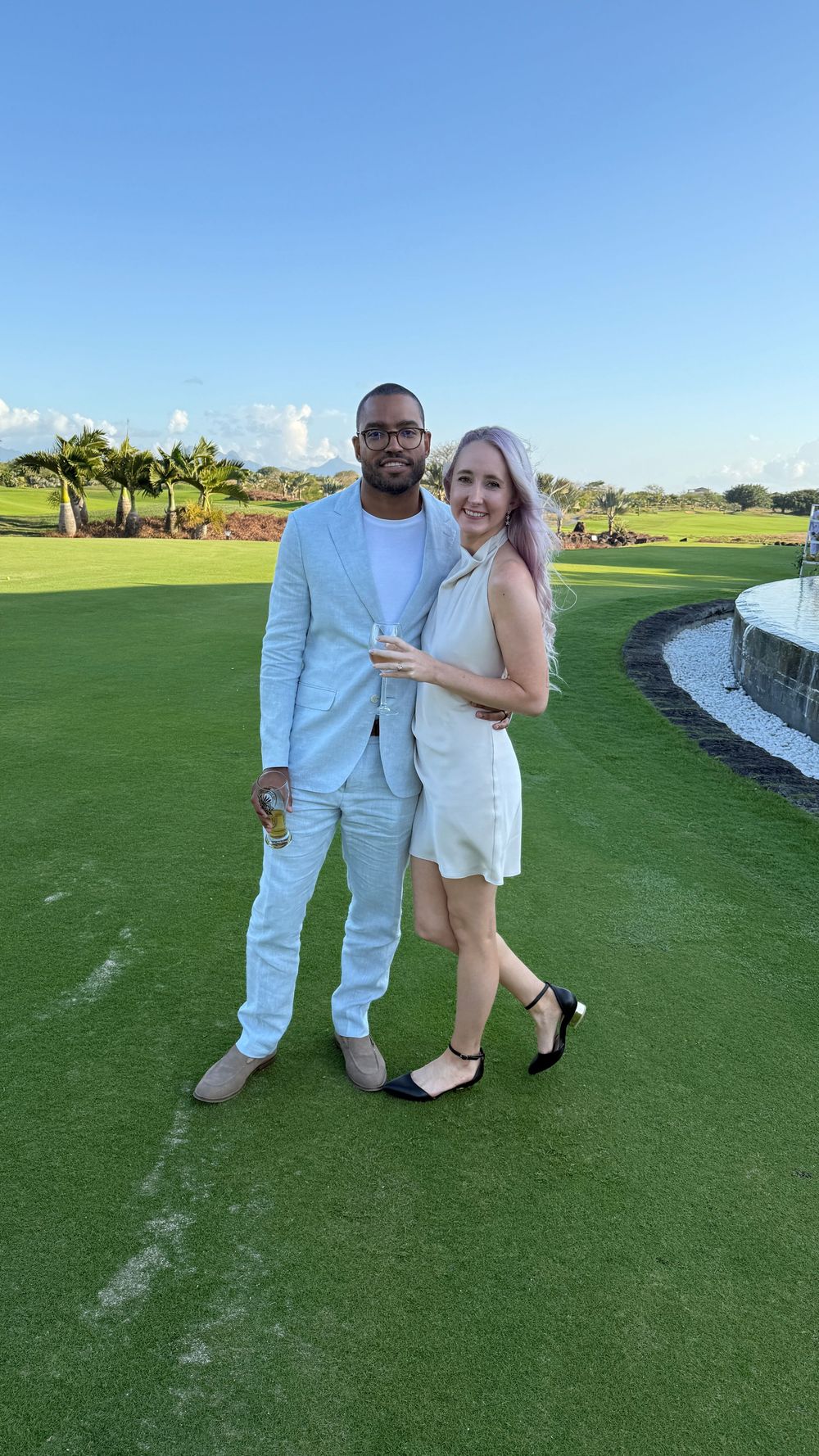 A couple poses together on a lush green golf course with blue skies and palm trees in the background.