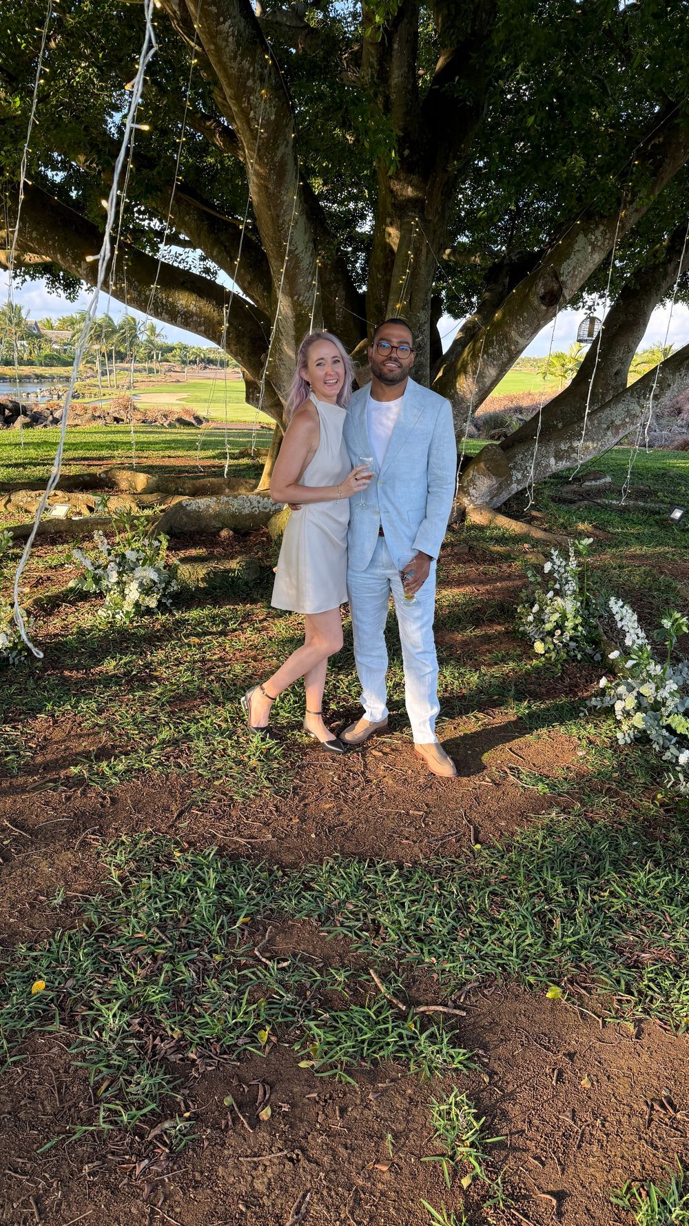 A couple poses together under a large tree decorated with hanging lights at an outdoor garden venue.