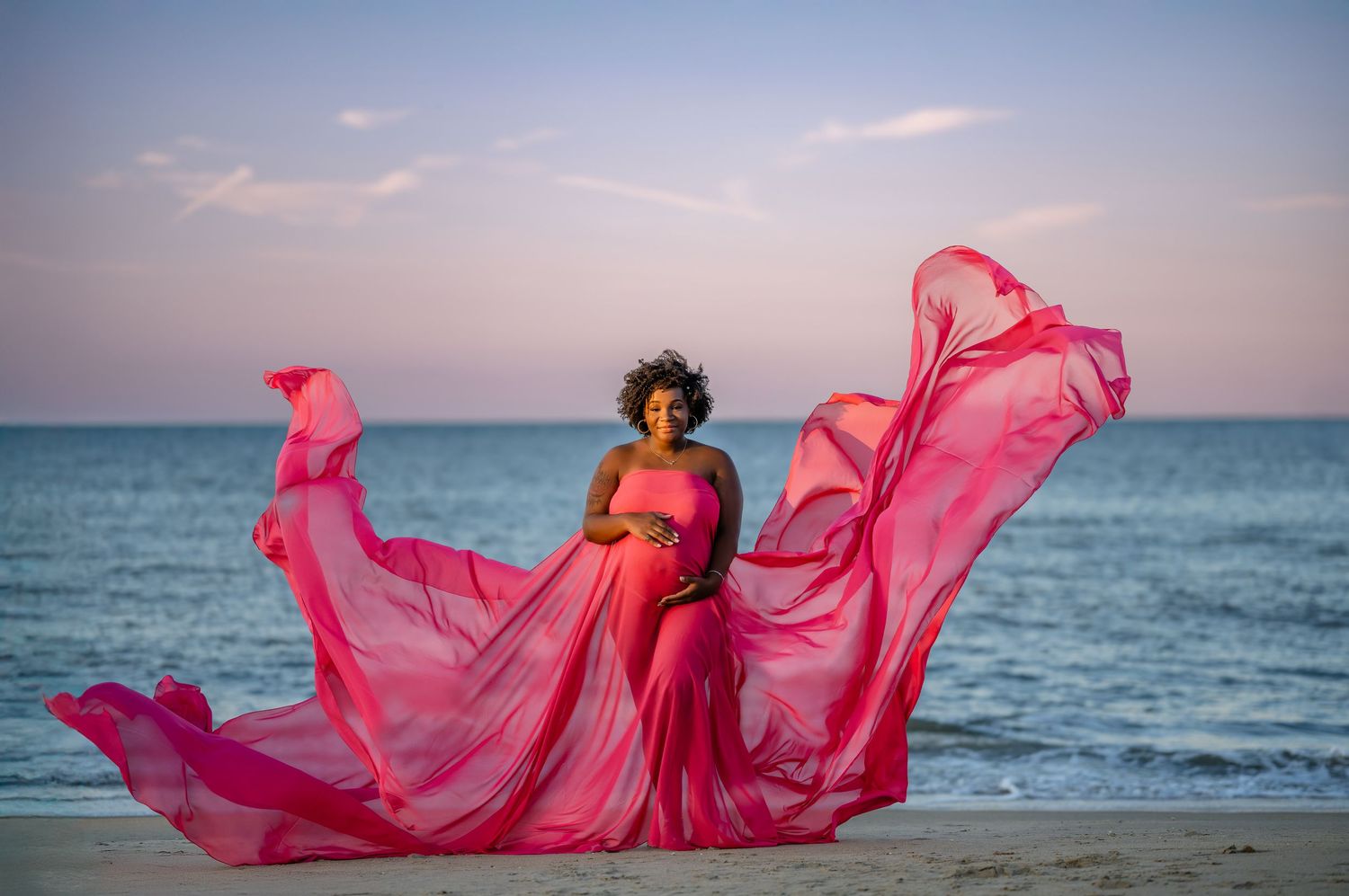 Maternity session in a flowing pink dress billows dramatically against an ocean backdrop at sunset, creating an ethereal beach scene.