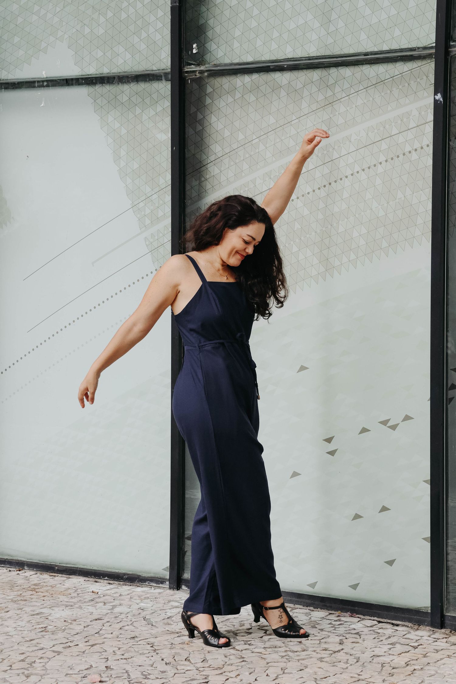 A woman in a navy jumpsuit and heels poses gracefully with one arm raised against a glass wall outdoors.
