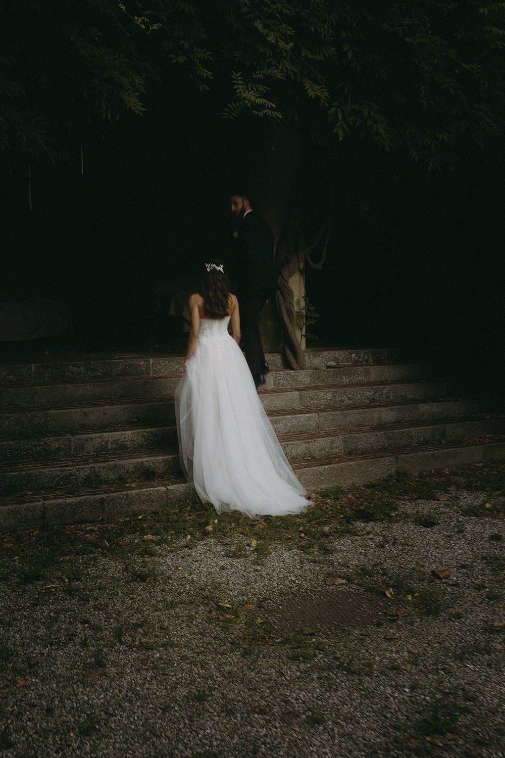 A bride in a flowing white wedding gown walks up dark stone steps in a moody forest setting.