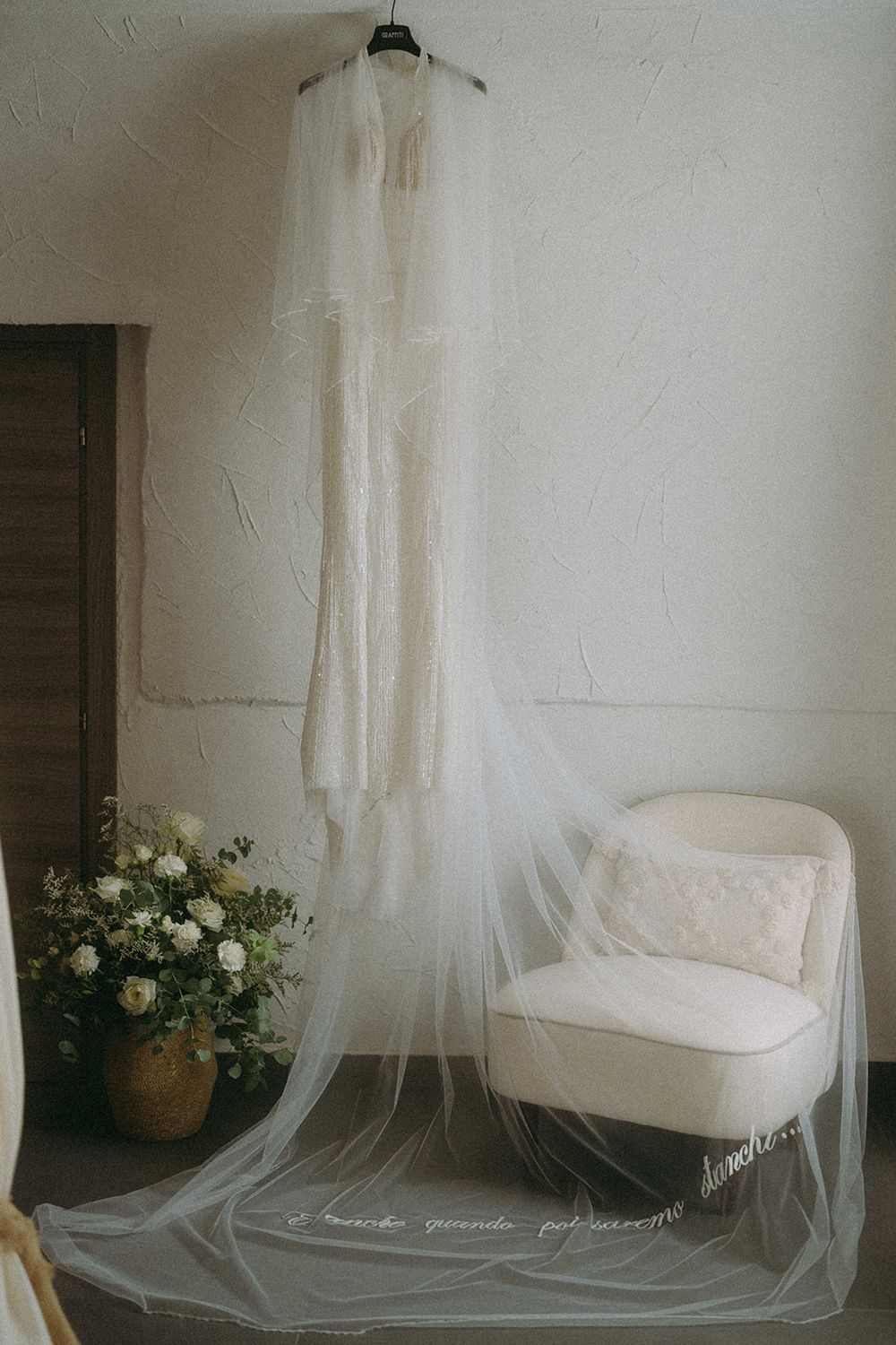 A delicate white wedding veil hangs in front of a white chair with white roses in a rustic interior setting.