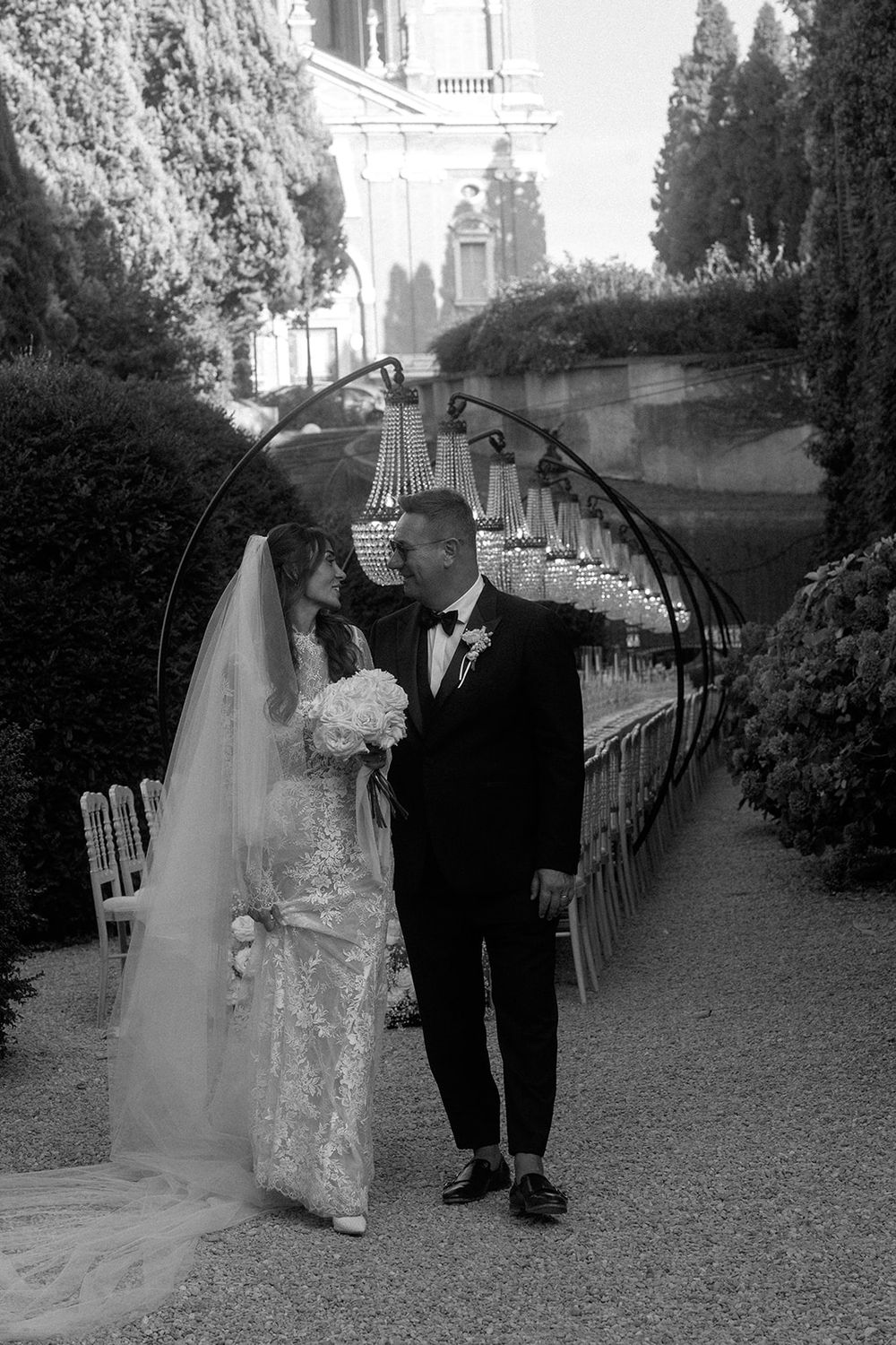 A couple walks beneath an arched garden pathway decorated with greenery.