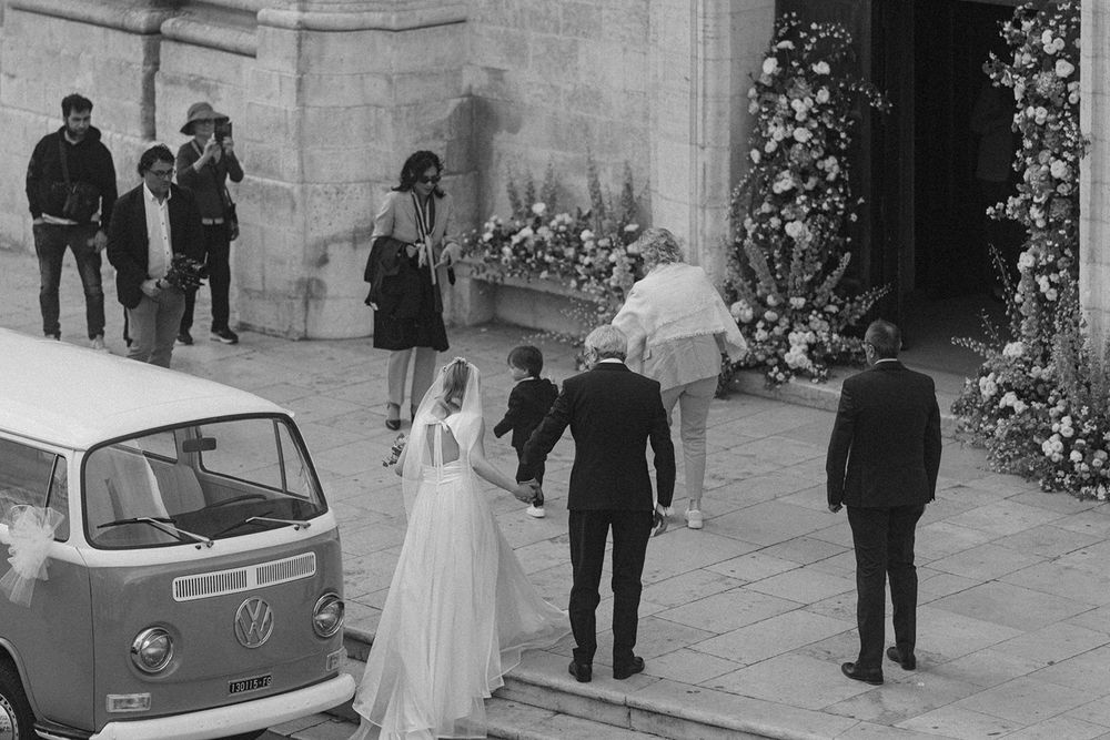 Wedding party exits stone church building with vintage car parked outside on cobblestone street.