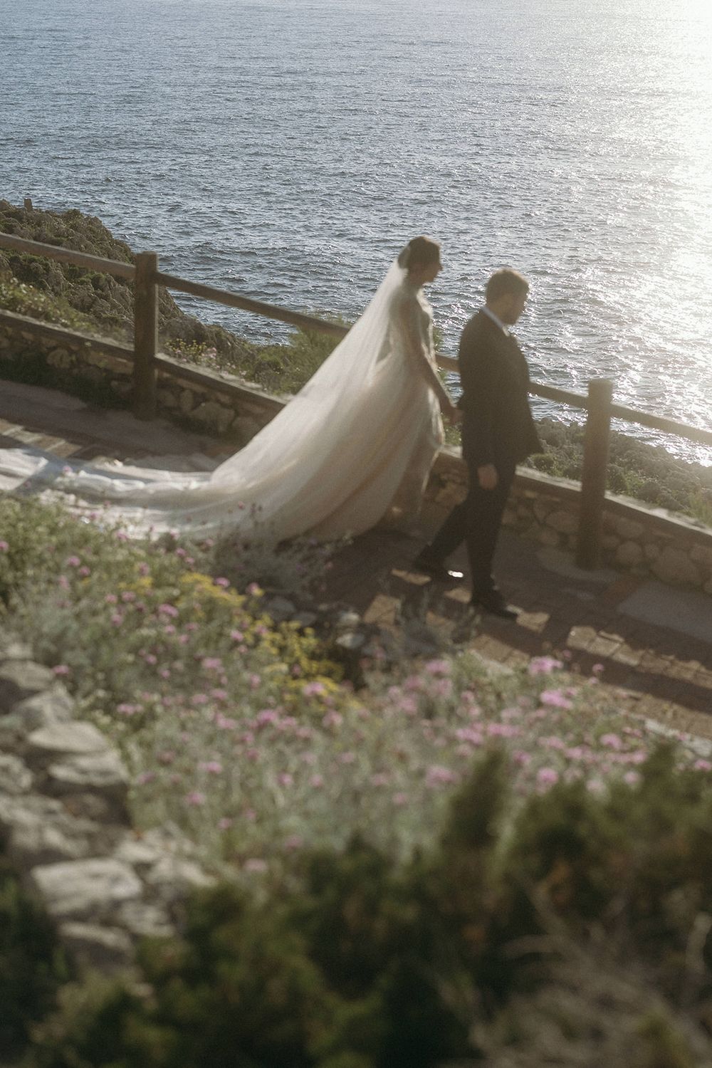 A couple in wedding attire stands by a coastal railing with flowing veil catching the ocean breeze at sunset.