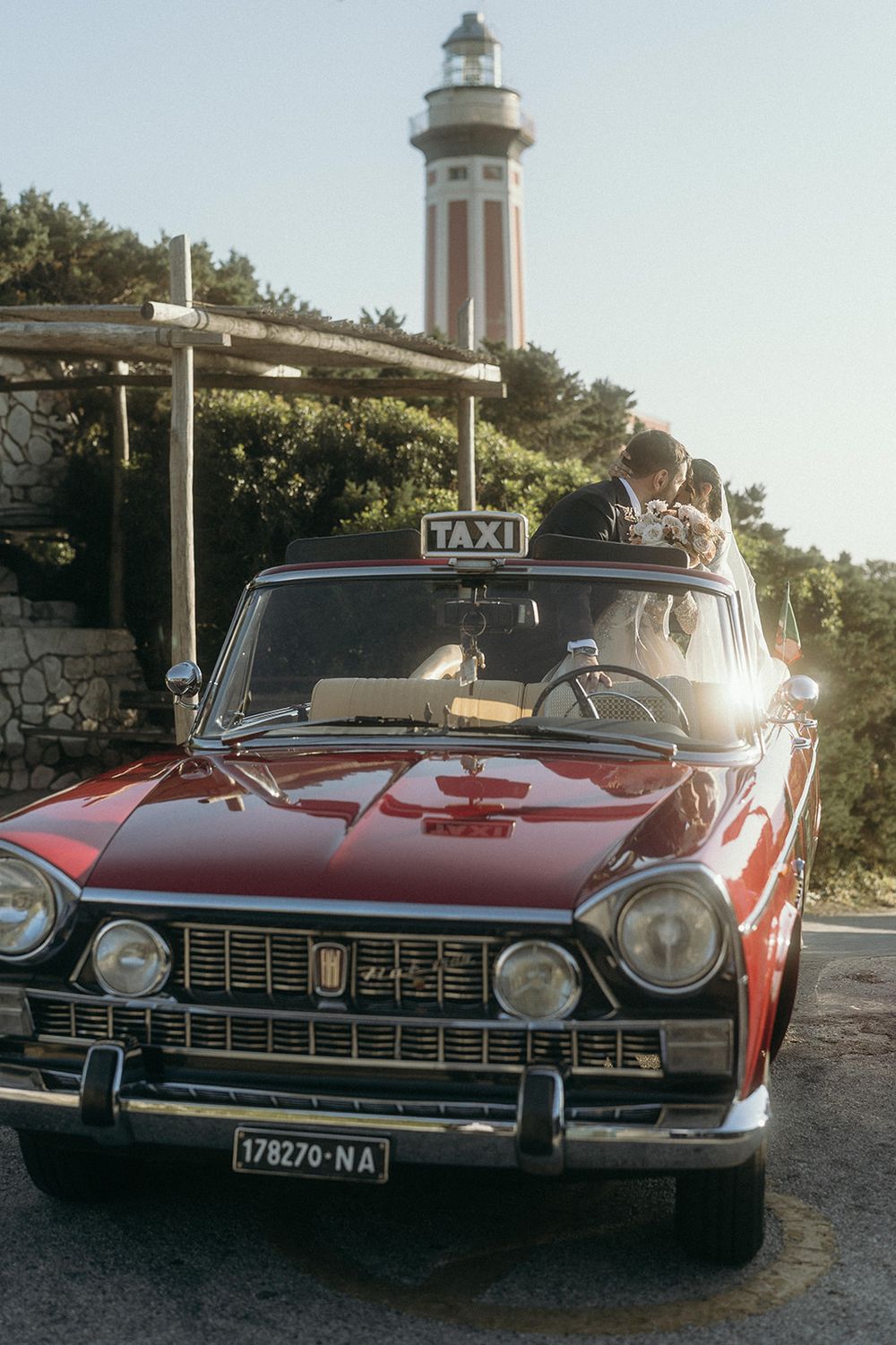 Vintage red and black taxi car parked near a lighthouse with sunlight shining through the trees.