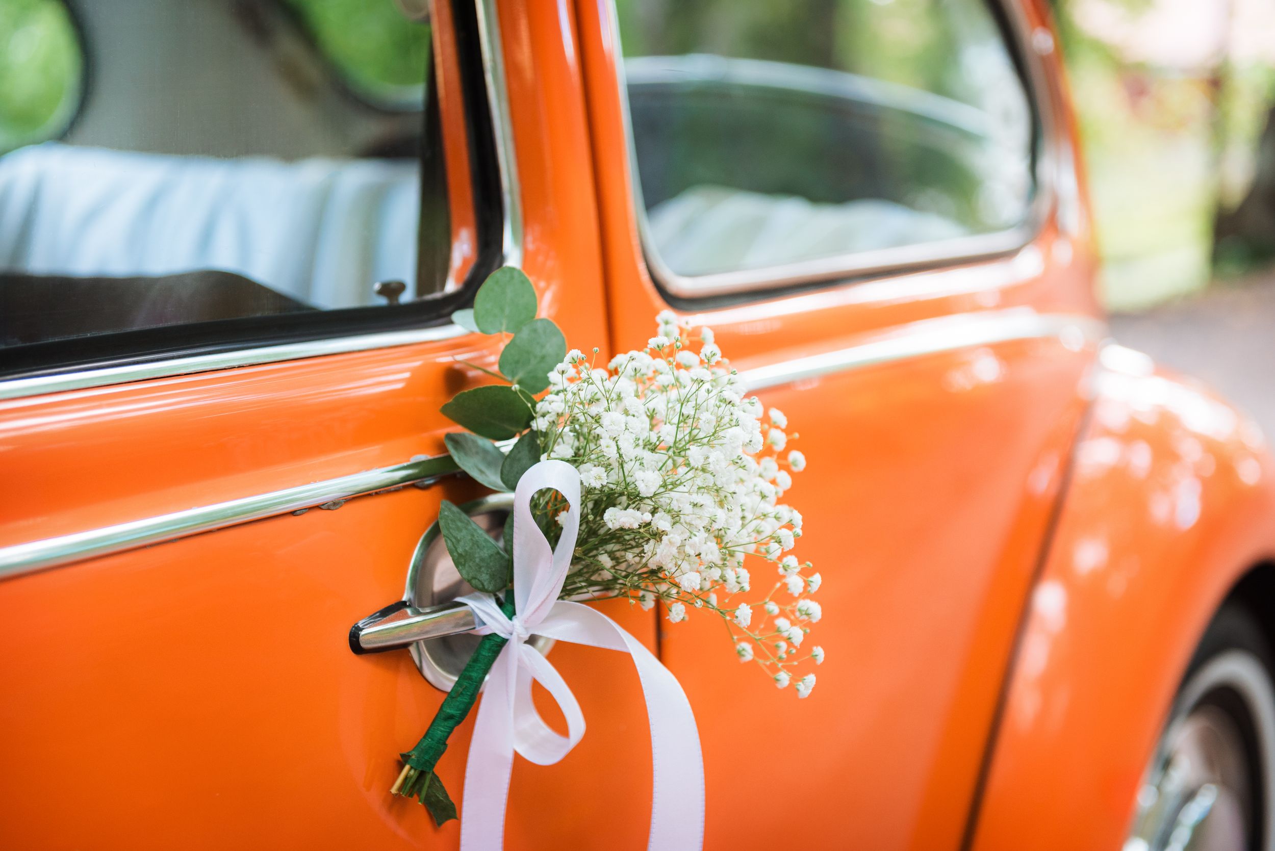 Oranssi hääauto, jonka kahvaa koristaa vaalea kukkakimppu, jossa on valkoinen rusetti.