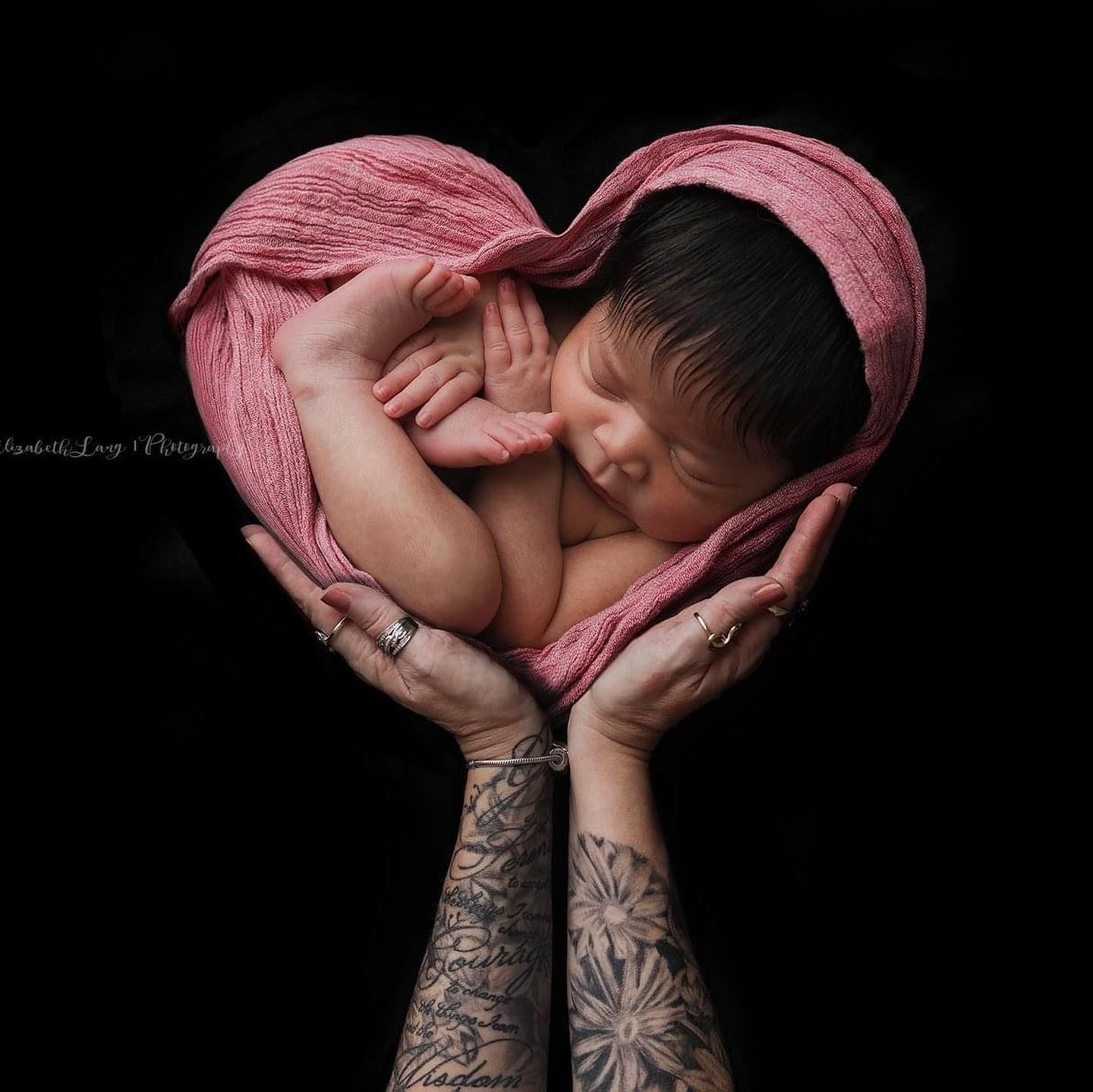 Tattooed arms cradle a newborn baby wrapped in soft pink fabric against a black background.