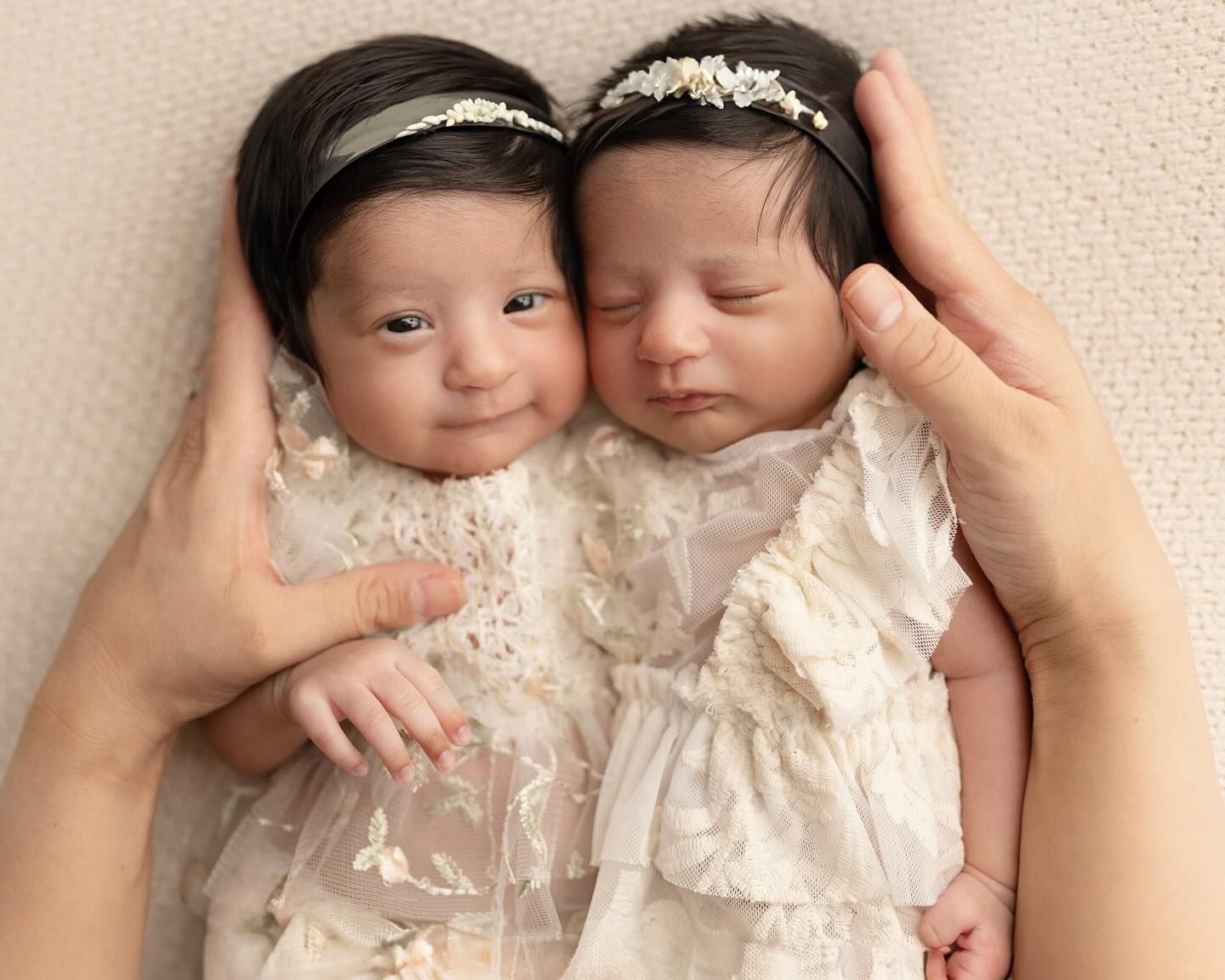 Two babies in white lace dresses lie close together on a beige textured background.