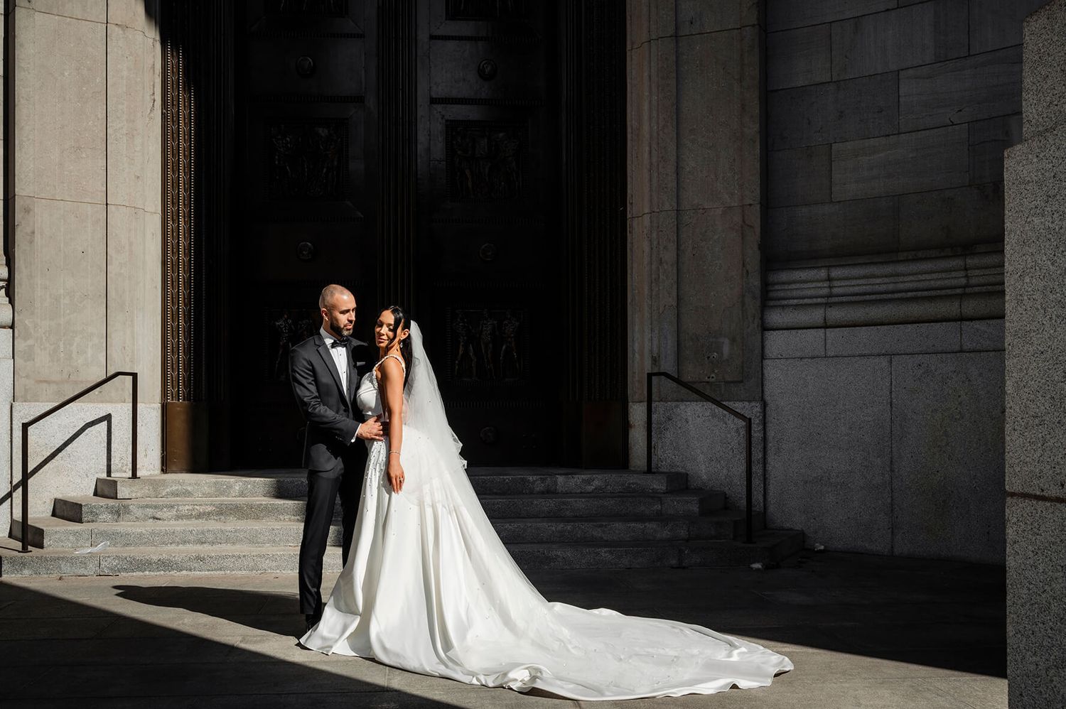 Elegant wedding couple posing on grand stone steps with dramatic shadows.
