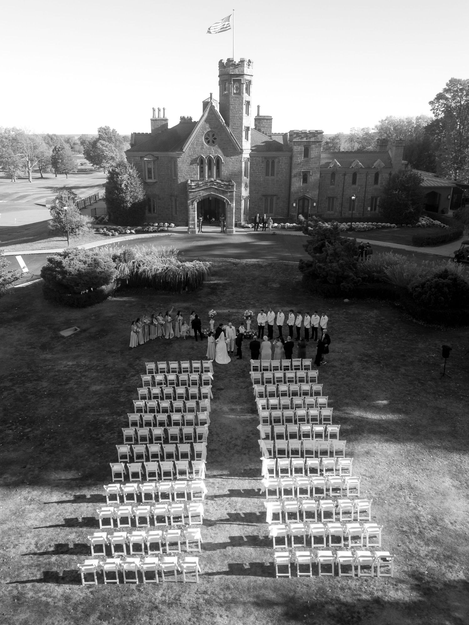 Aerial view of an outdoor wedding ceremony setup with white chairs facing a Gothic castle venue during autumn.