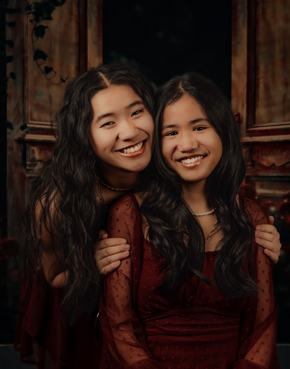 Two people wearing burgundy lace dresses pose together in front of a dark wooden backdrop.