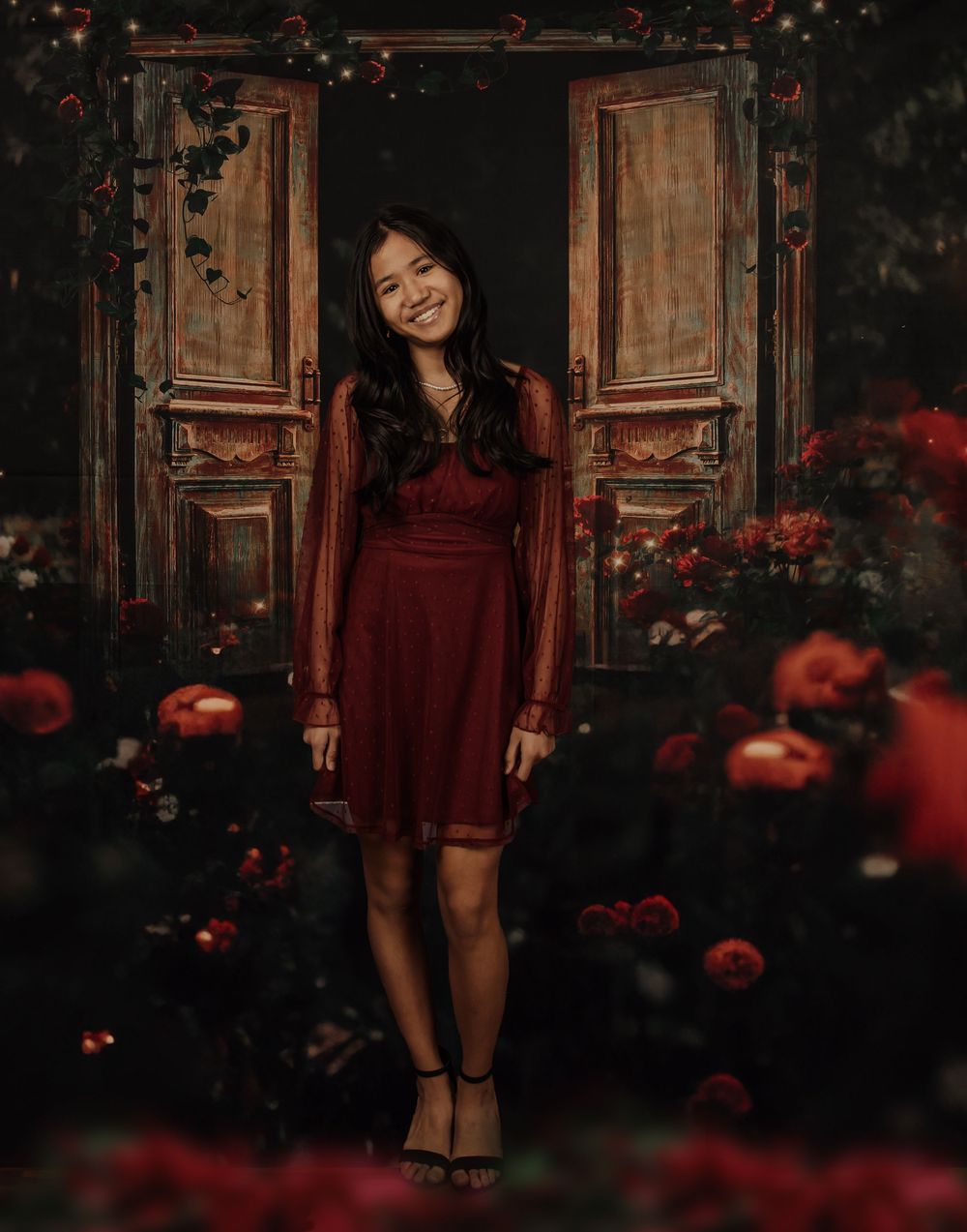 A burgundy dress stands out against a rustic wooden door backdrop with red flowers in a moody garden setting.