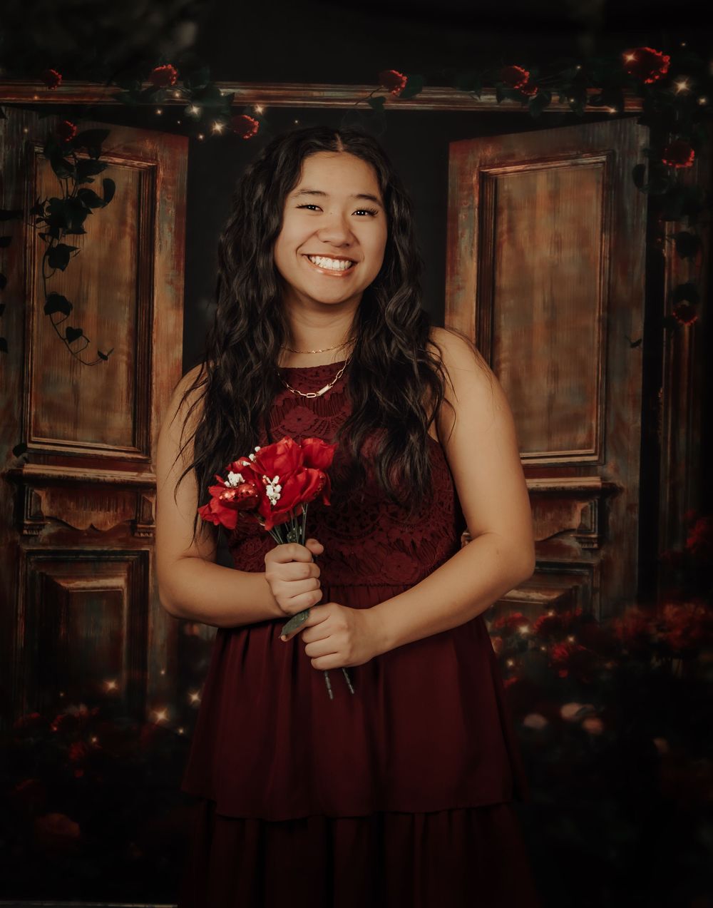 Young girl in burgundy dress poses elegantly against rustic wooden backdrop with floral accents.