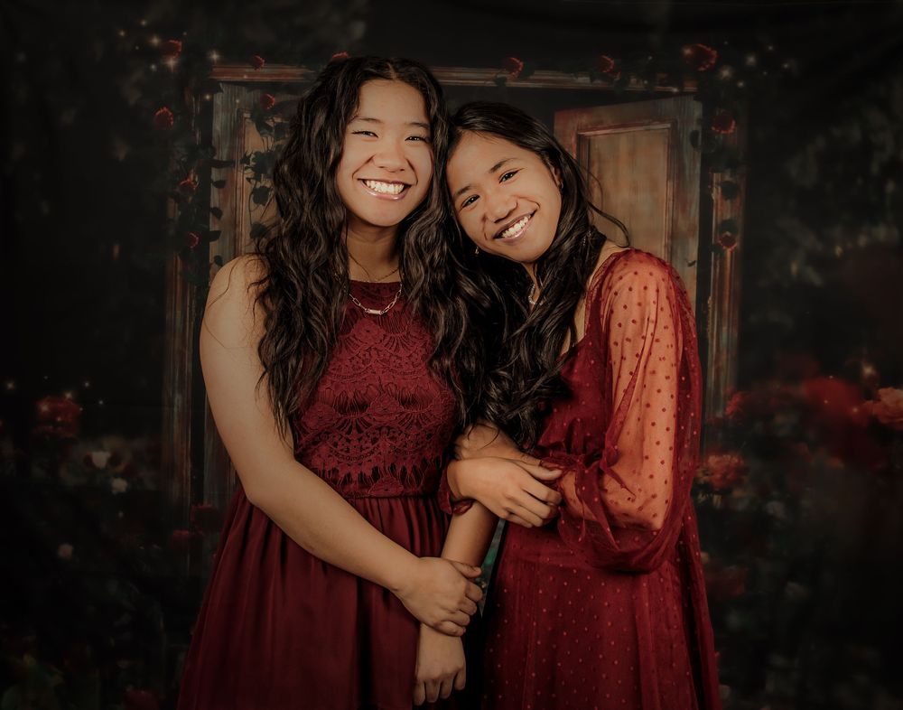 Two people in burgundy dresses share a joyful moment together against a dark rustic backdrop.