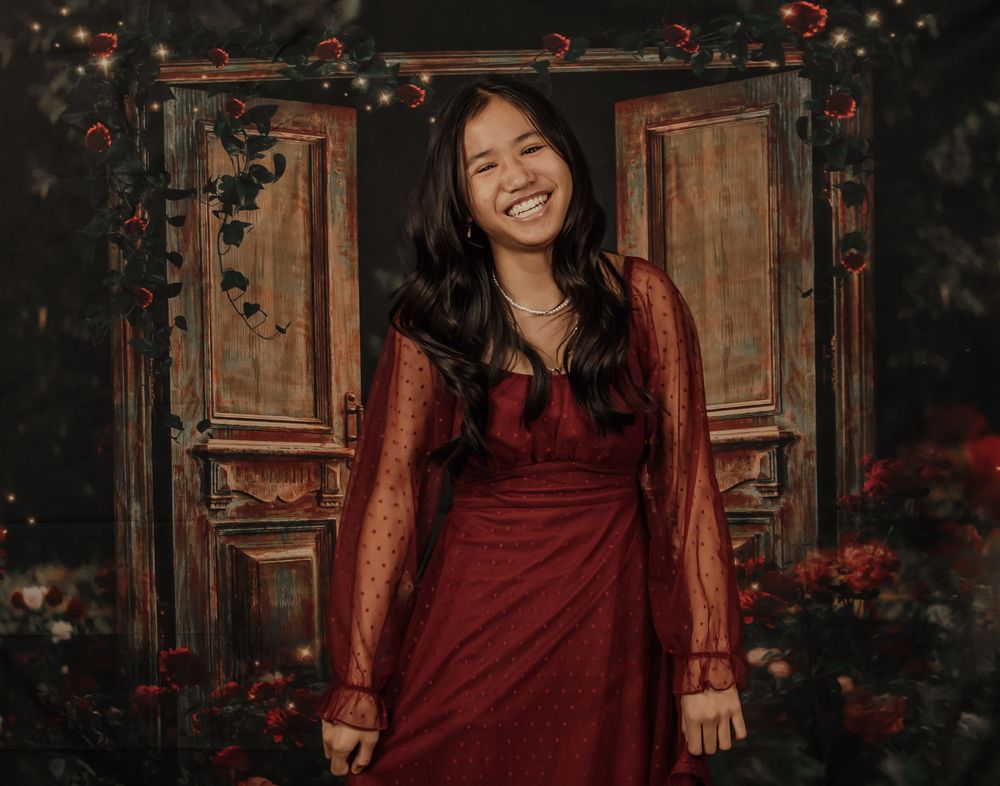 A person in a burgundy dress poses against a rustic wooden door with red flowers in a moody photoshoot setting.