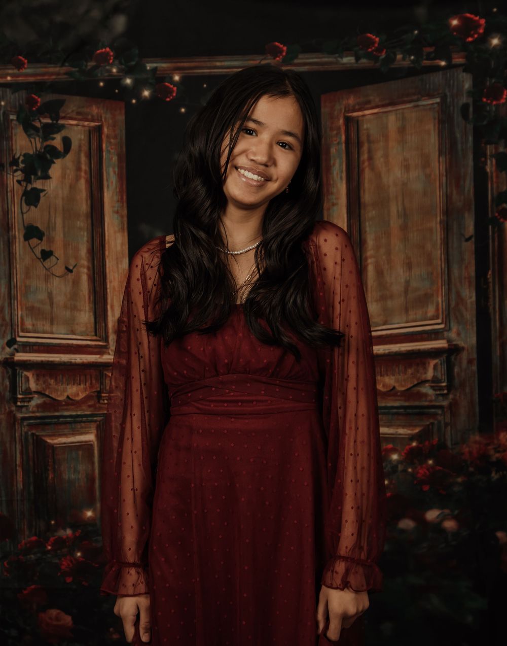 Woman in burgundy lace dress poses against rustic wooden backdrop with moody lighting and floral accents.