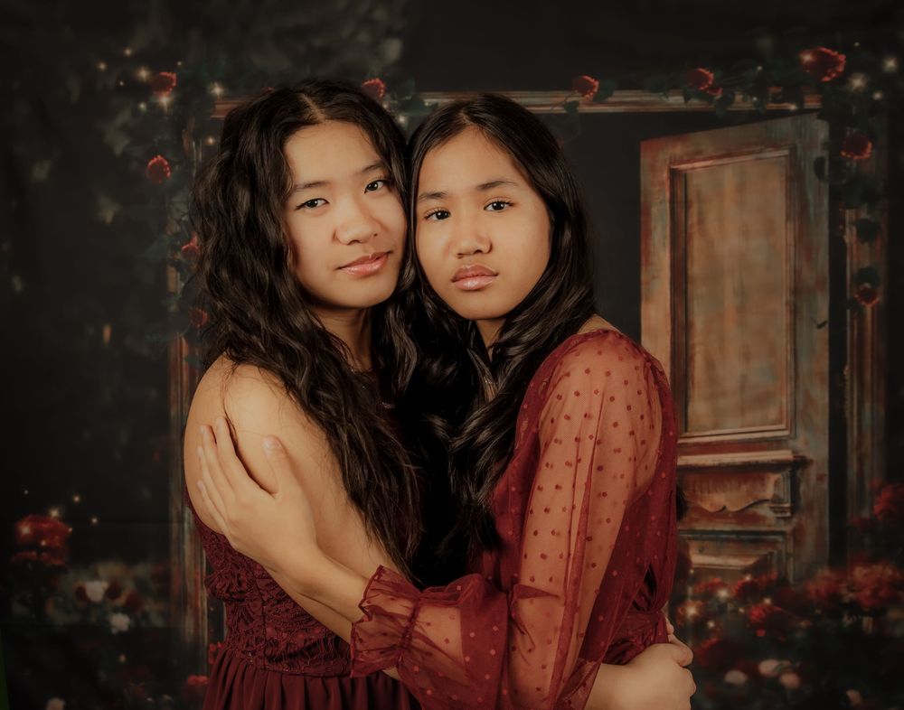 Two people in burgundy dresses embrace in a romantic photoshoot with rustic wooden backdrop and soft lighting.