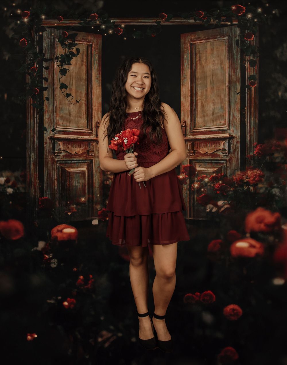 Dark moody portrait session with burgundy dress against vintage wooden door and red floral accents.