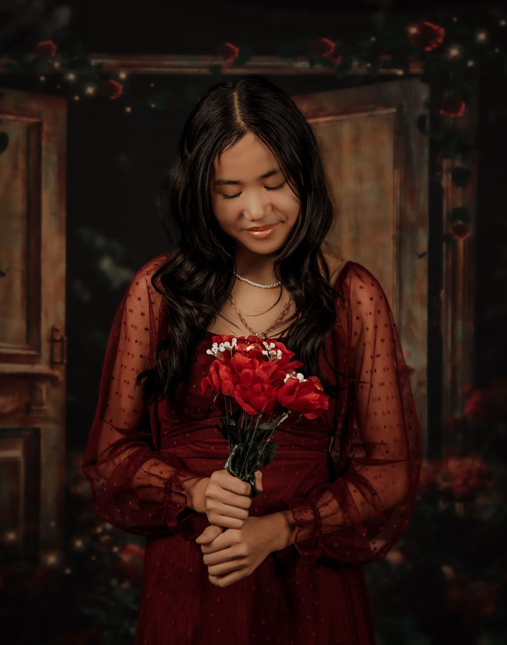 Person in a burgundy lace dress holds red roses while looking down in a moody portrait setting.