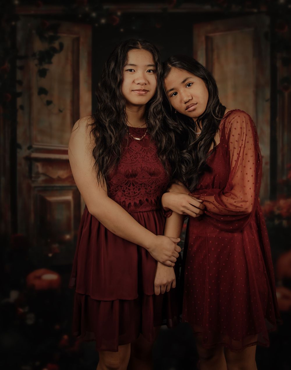 Two children in burgundy dresses pose together in a moody indoor portrait session.