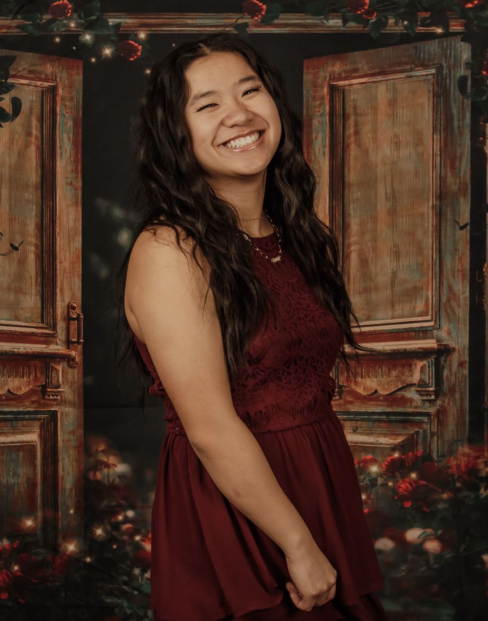 A woman in a burgundy lace dress poses against a rustic wooden backdrop with red floral accents.