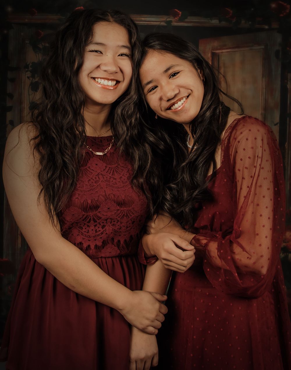 Two friends wearing burgundy dresses share a joyful moment together in a warm-toned portrait.
