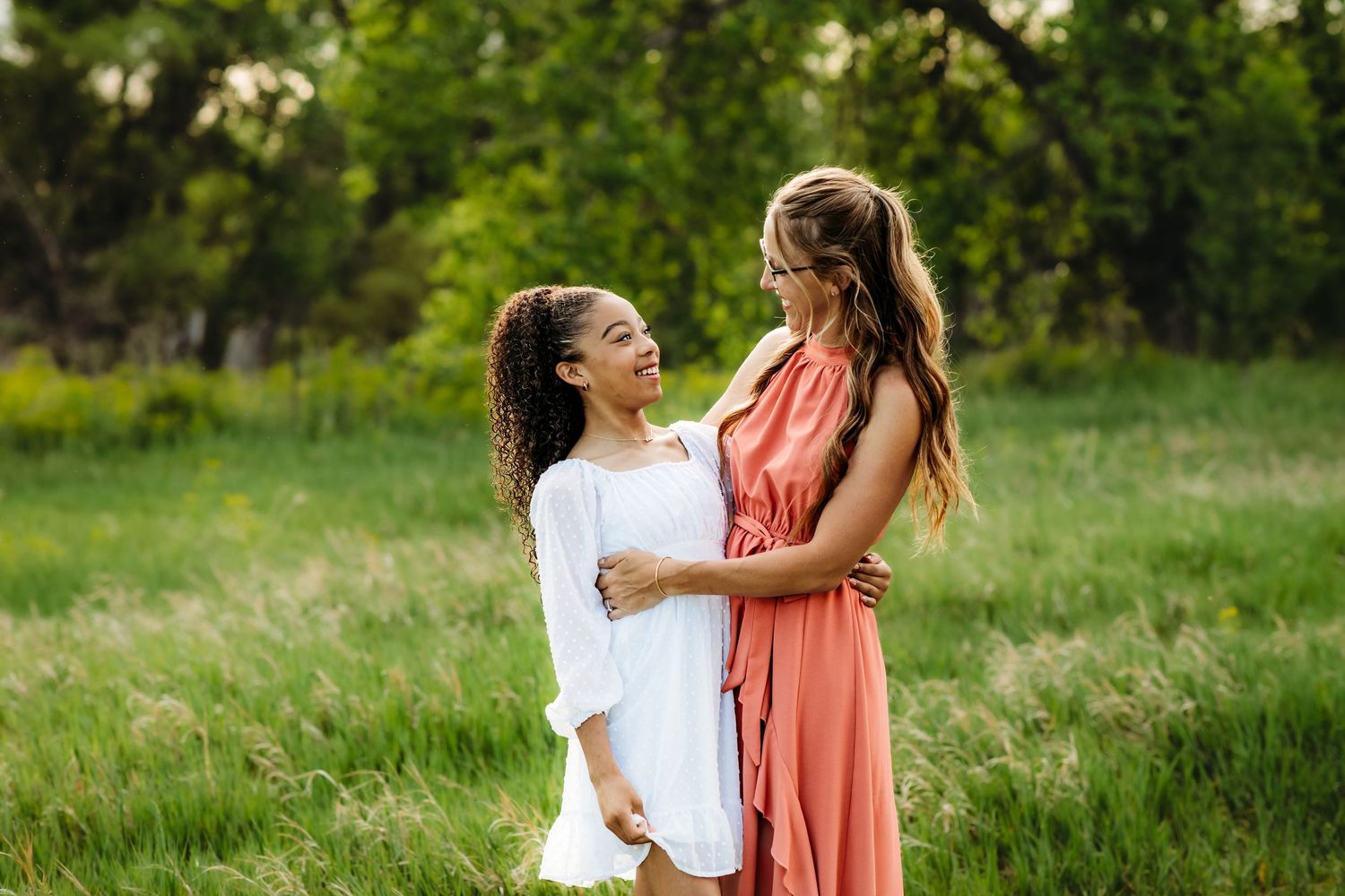 A couple in white and coral outfits share tender moments while walking through a sunlit grassy meadow.