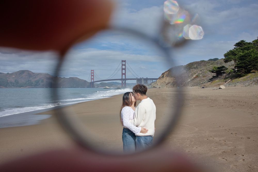 Couple framed with engagement ring walking along Baker Beach with the iconic Golden Gate Bridge visible in the background on a cloudy day.