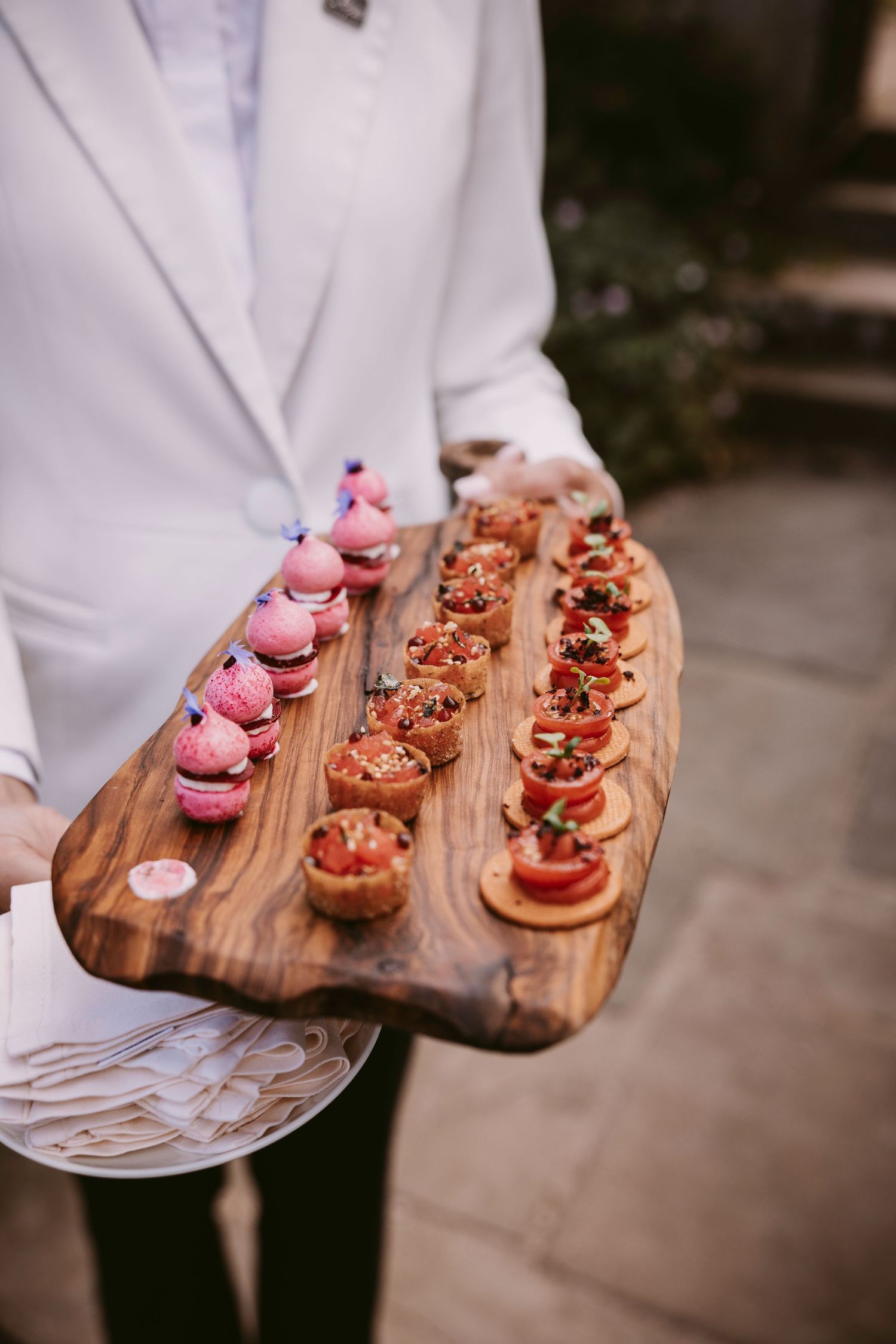 A wooden serving board displays an elegant arrangement of pink macarons and bite-sized dessert canapes.