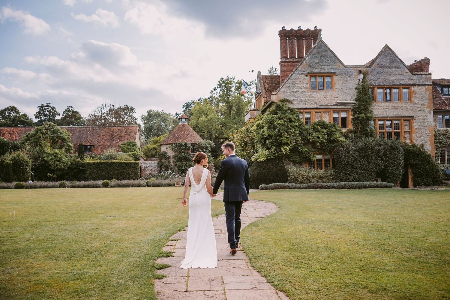 A couple walks together on a grassy lawn with a grand brick building in the background during a sunny day.